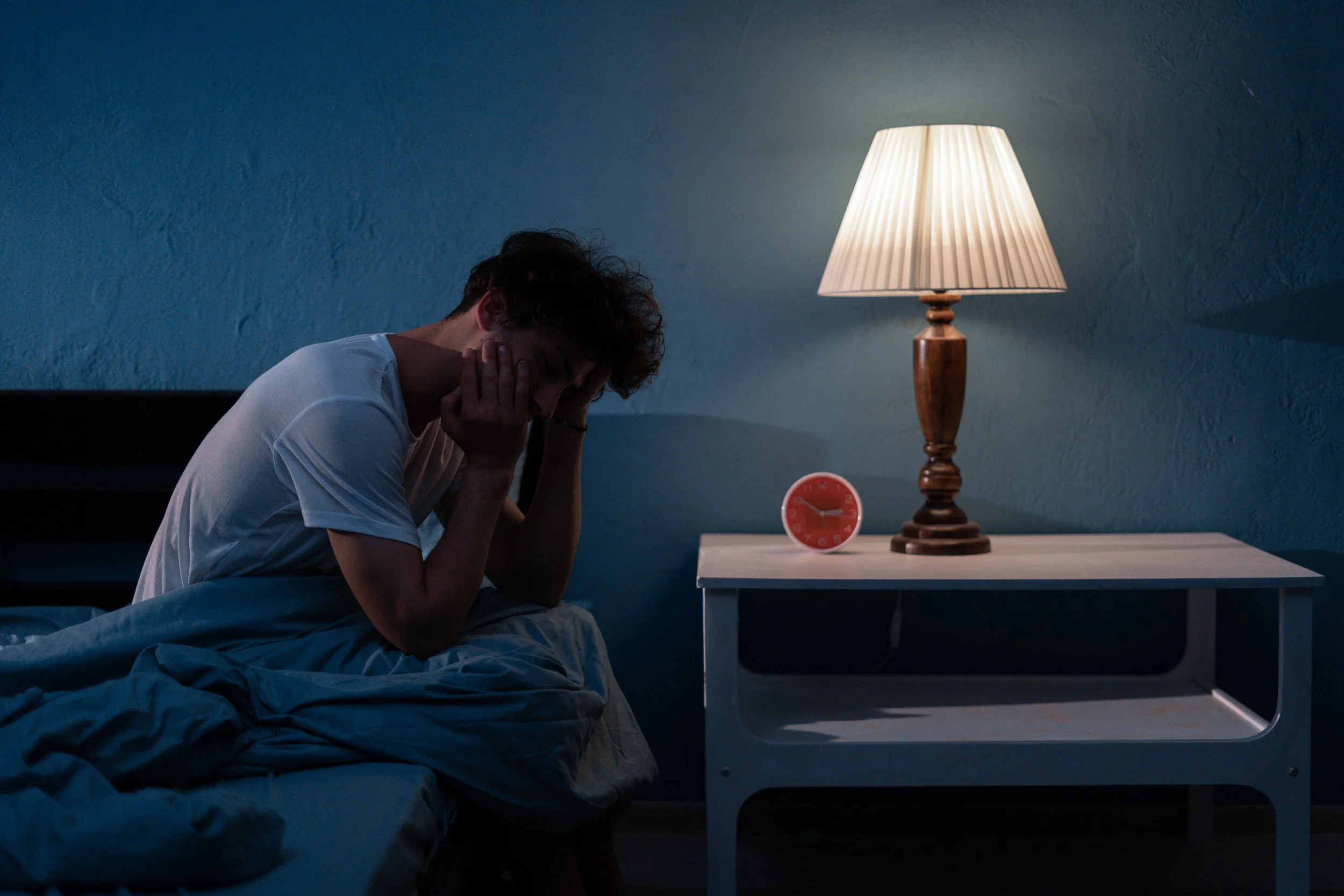 A young man sits on a bed, holding his head in his hands, appearing distressed. A bedside table next to him has a lit lamp and a red clock showing 3:05.