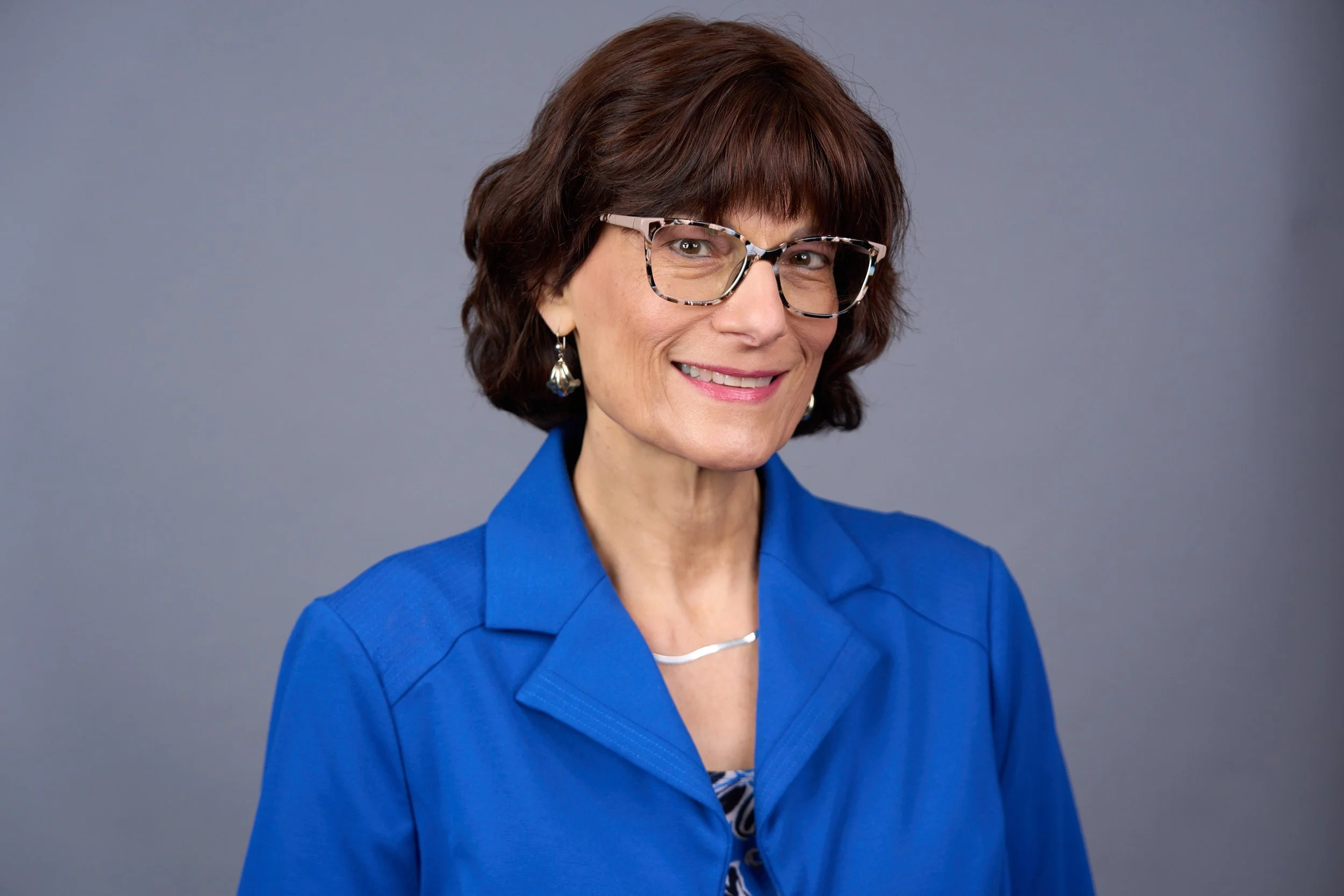 Portrait of a smiling woman with short brown hair, wearing glasses, earrings, a blue blazer, and a patterned blouse against a plain gray background.