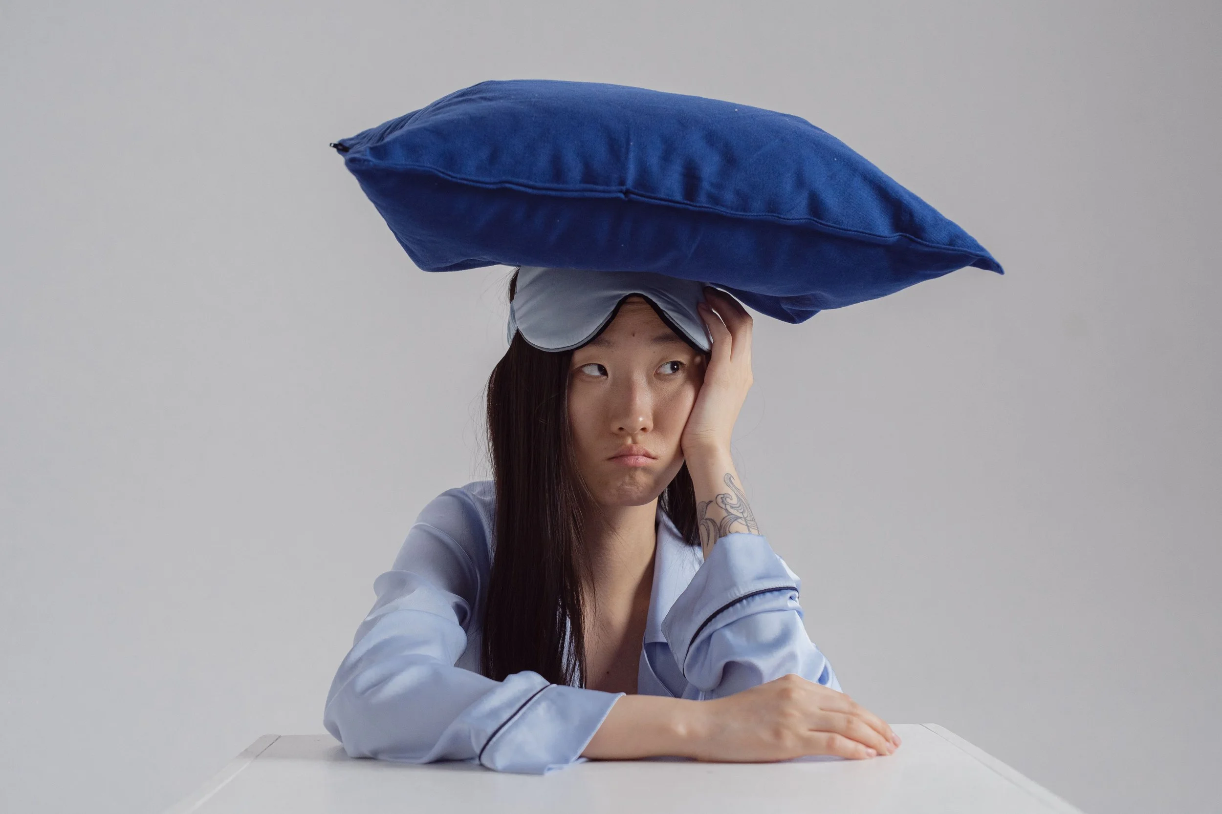 A woman with long dark hair, wearing a light blue shirt and goggles, balances a large blue pillow on her head while resting her chin on her hand at a white table against a plain gray background.