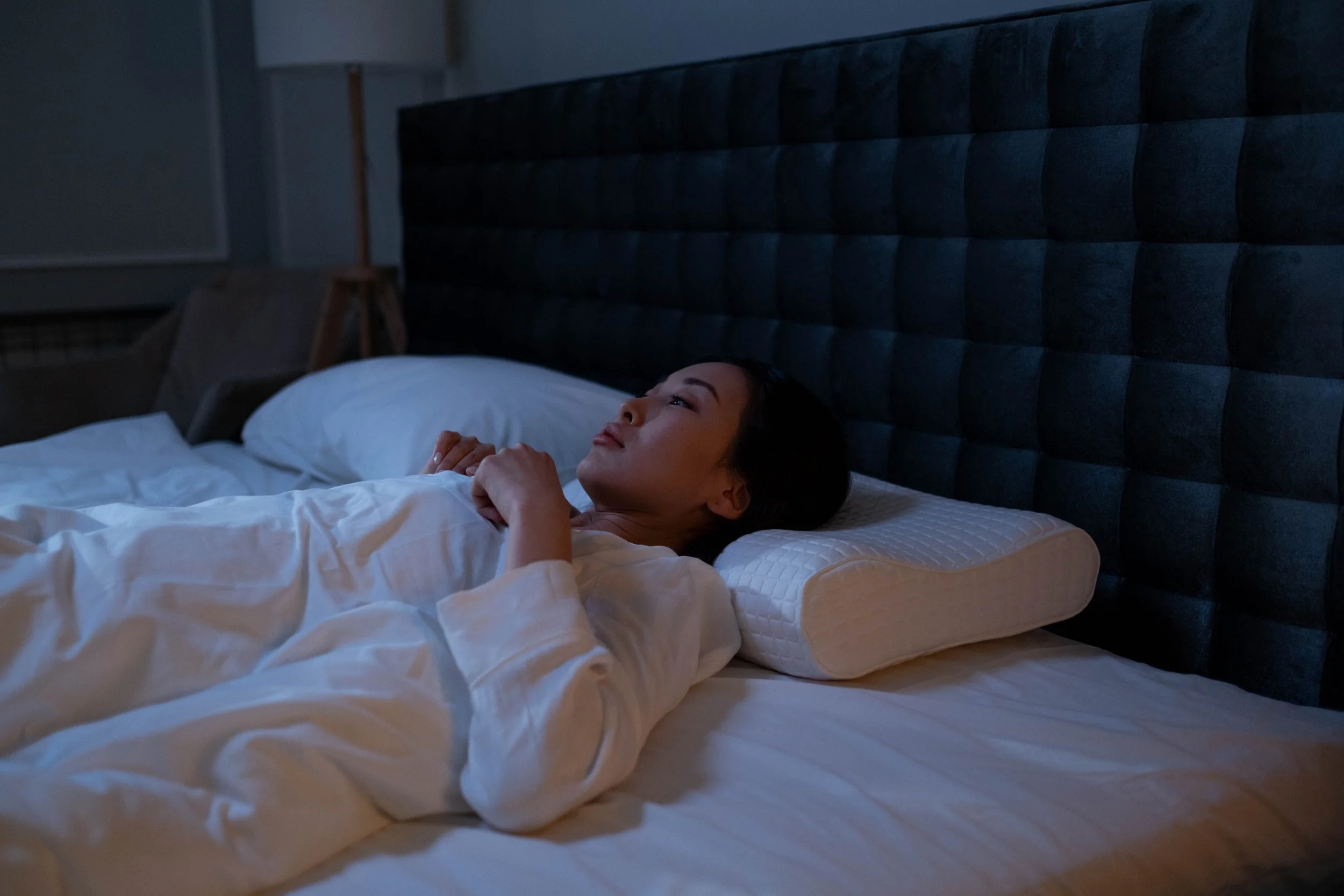 A woman lying in bed at night, looking peaceful, with a dark headboard behind her and her head resting on a white pillow.