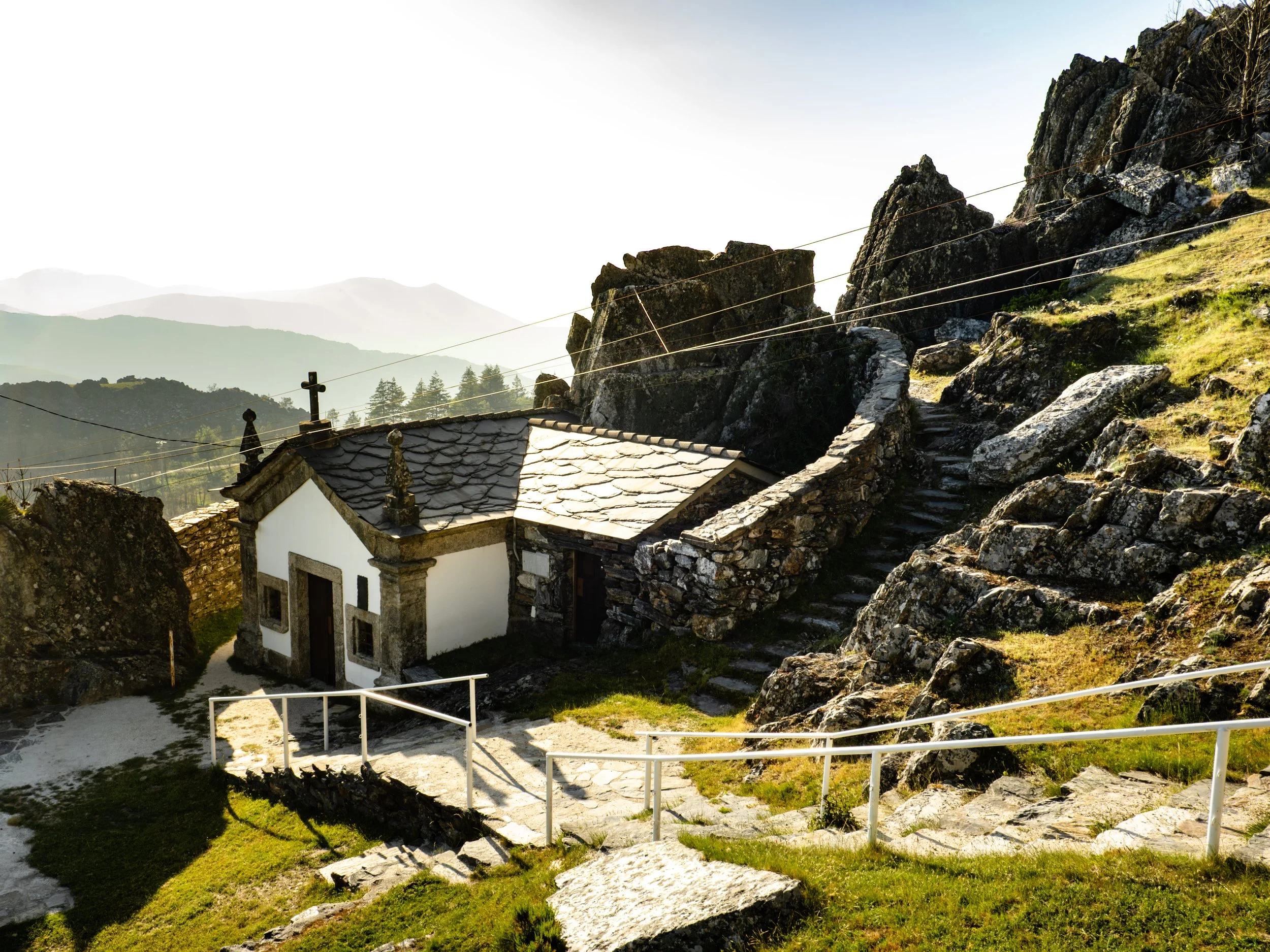Casa branca de telhado de ardósia em meio a uma paisagem montanhosa com rochas e vegetação verde, cercada por uma cerca de fios e escadaria de pedra.