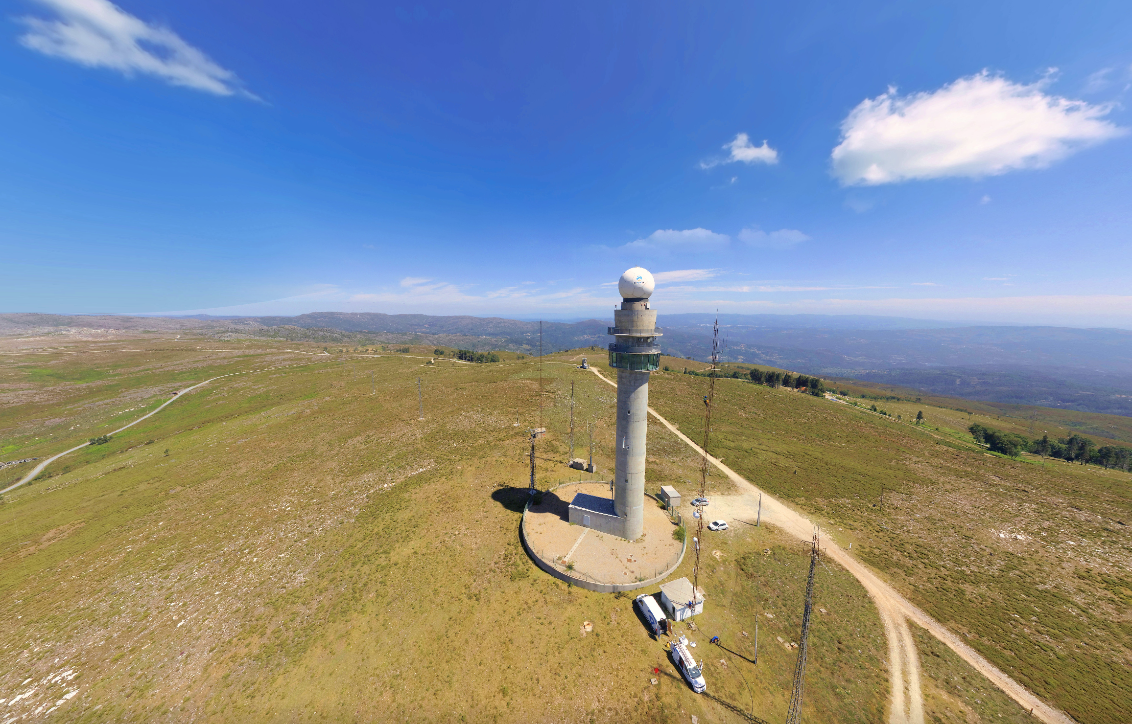 Imagem de uma torre de radar em uma área elevada com vegetação, estrada de terra ao redor, céu azul com algumas nuvens e horizonte de montanhas ao fundo.
