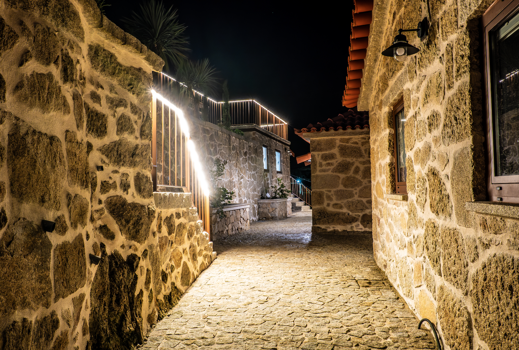 Rua de pedra entre casas de pedra à noite, iluminada por luzes ao longo da parede e luminárias das casas, com céu estrelado ao fundo.