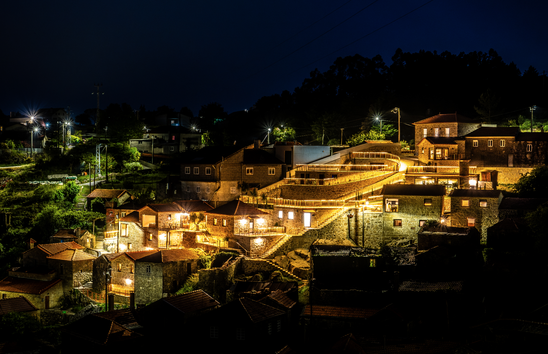 Cena noturna de uma cidade com casas de pedra e ruas iluminadas por luzes amareladas, rodeada por árvores e colinas sob céu escuro.