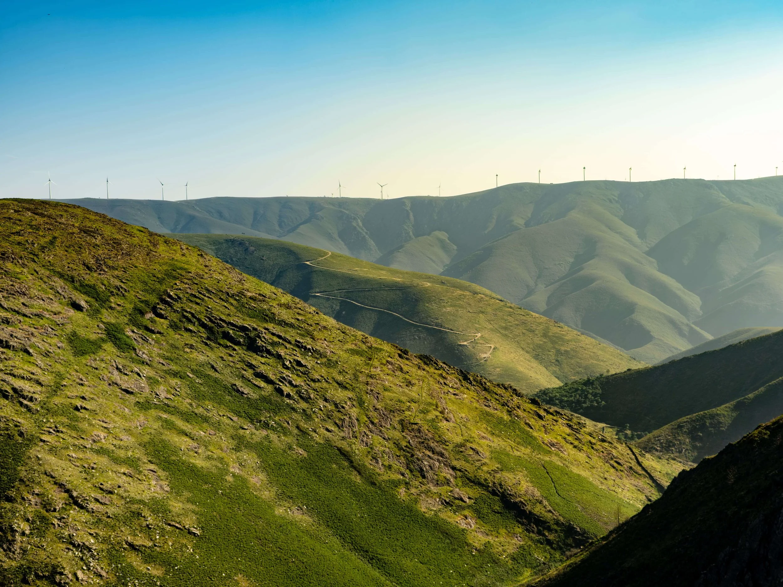 Cadeias de montanhas verdes sob céu claro com painéis de energia eólica ao longo do topo.