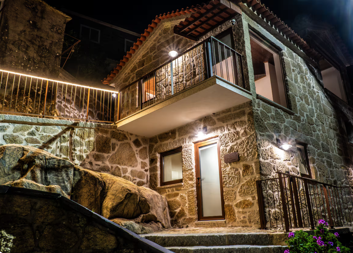 Casa de pedra iluminada à noite, com varanda e escadas na frente, flores no jardim.