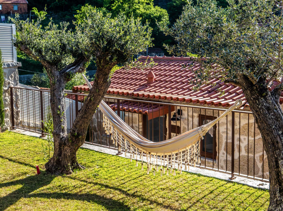Hamac pendurado entre duas árvores no jardim de uma casa com telhado de telhas vermelhas.