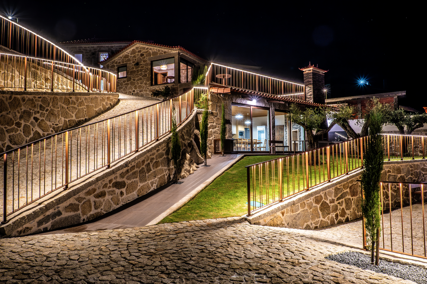 Casa de pedra iluminada à noite, com escadas, varanda, árvores e quintal com iluminação decorativa.