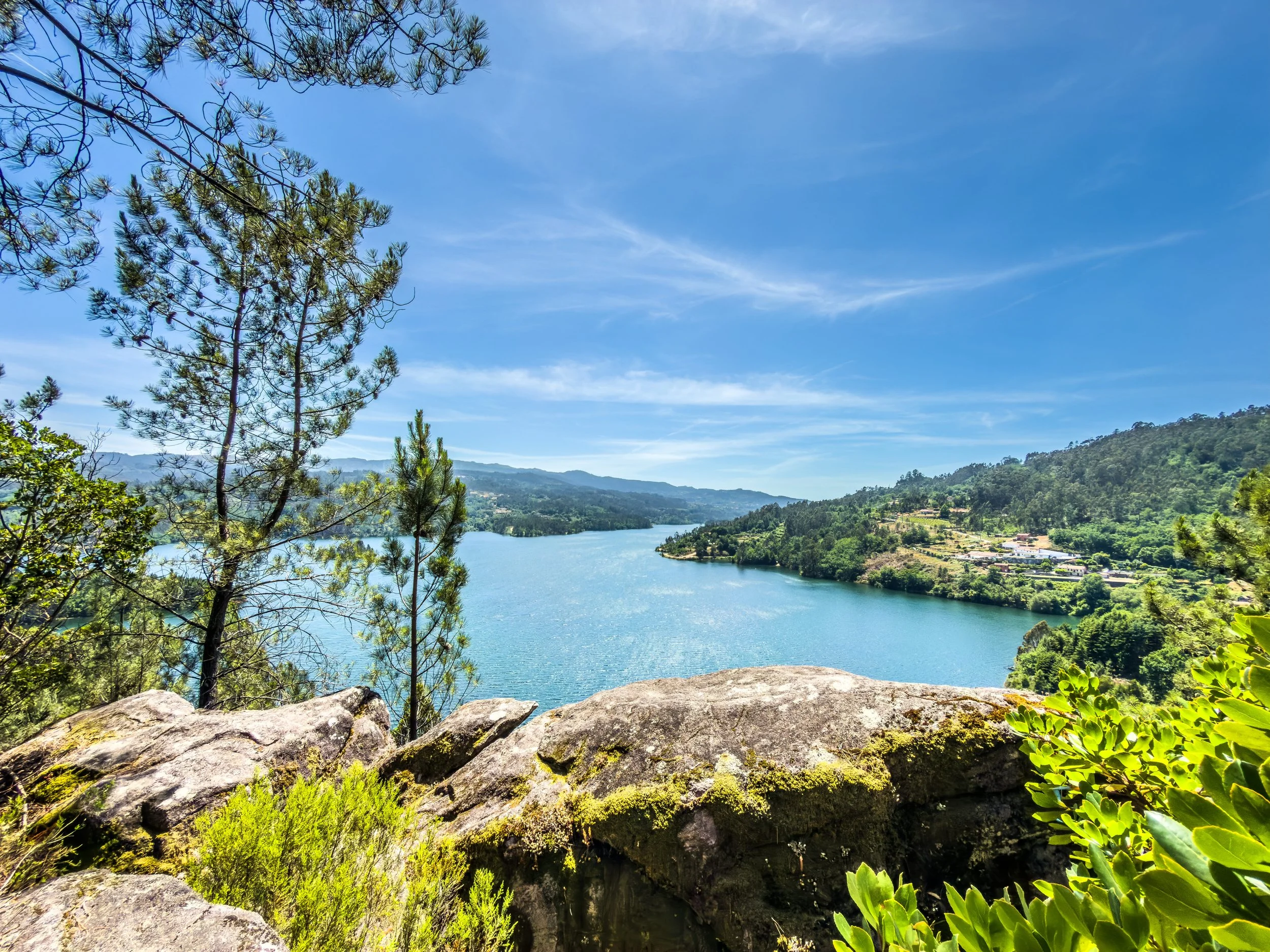Vista de um rio cercado por montanhas verdes, árvores ao redor, céu azul com nuvens finas, vista de um ponto de observação com pedras na frente.