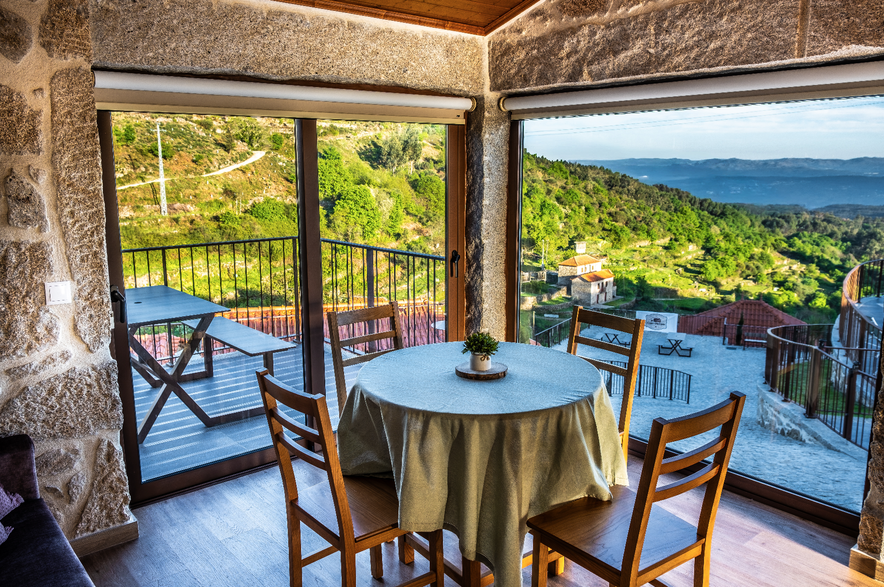 Sala de jantar com mesa redonda coberta por toalha bege, cercada por cadeiras de madeira, com vista para uma varanda ao ar livre e uma paisagem de montanhas verdes ao fundo.