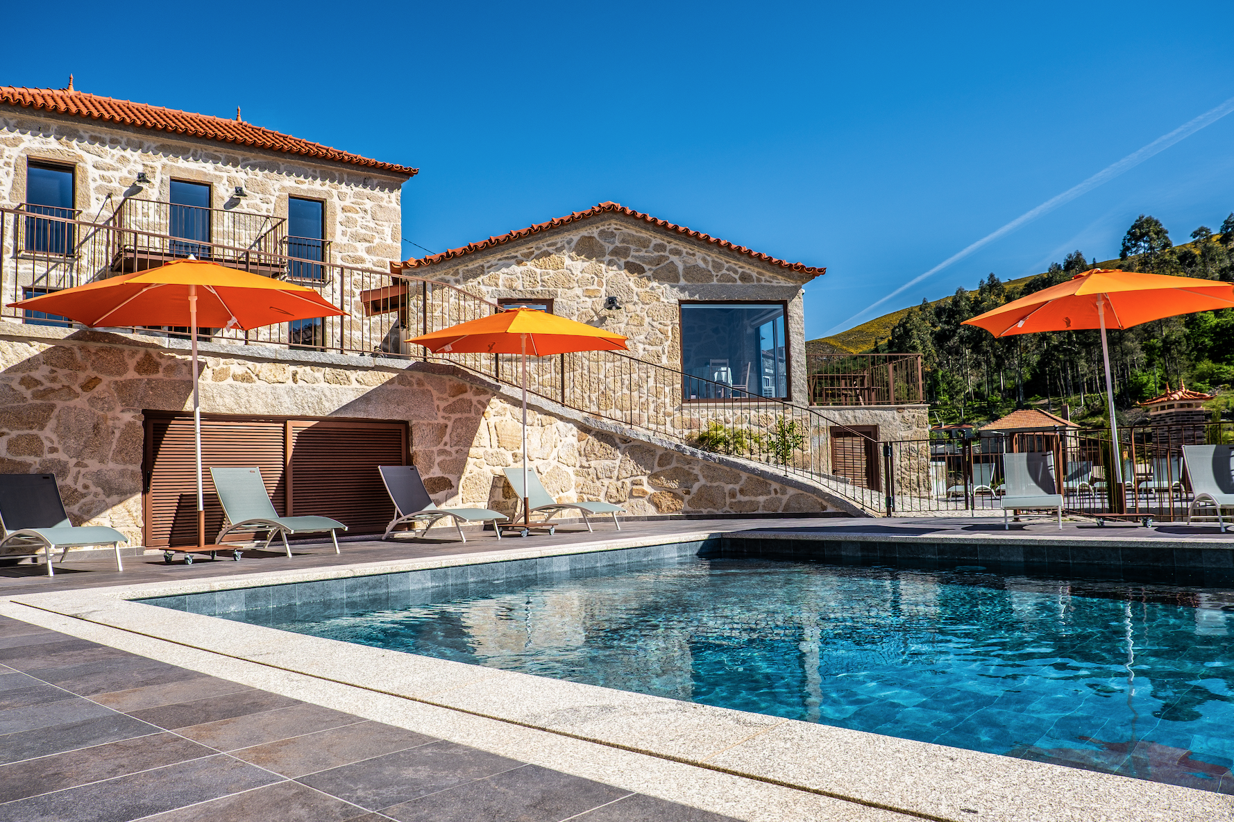 Piscina com espreguiçadeiras e guarda-sóis na área de lazer de uma residência de estilo rústico, com casas de pedra, janelas grandes e ambiente ao ar livre com montanhas ao fundo.