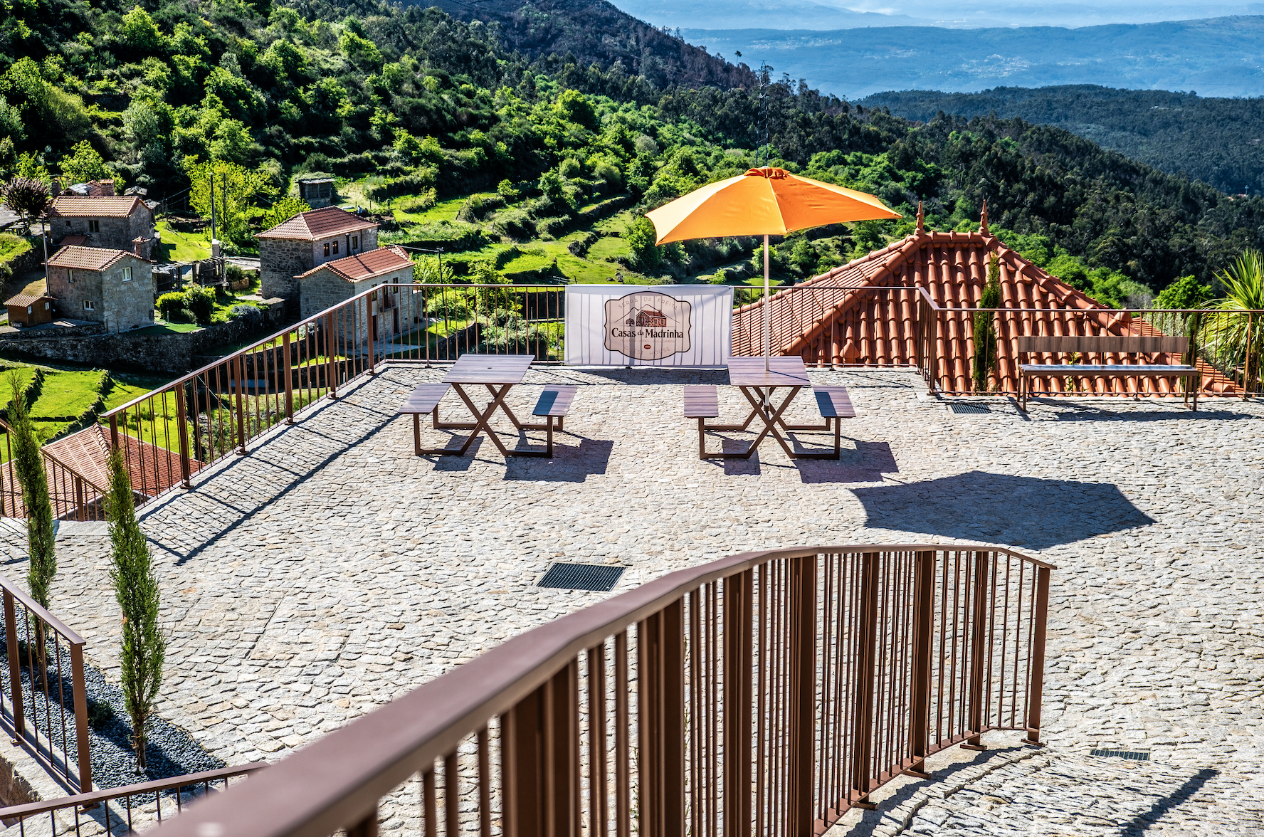 Vista de uma varanda com piso de pedras, cercada por uma grade de ferro, com duas mesas de madeira, uma sombra de guarda-sol laranja e uma vista de casas e montanhas verdes ao fundo.