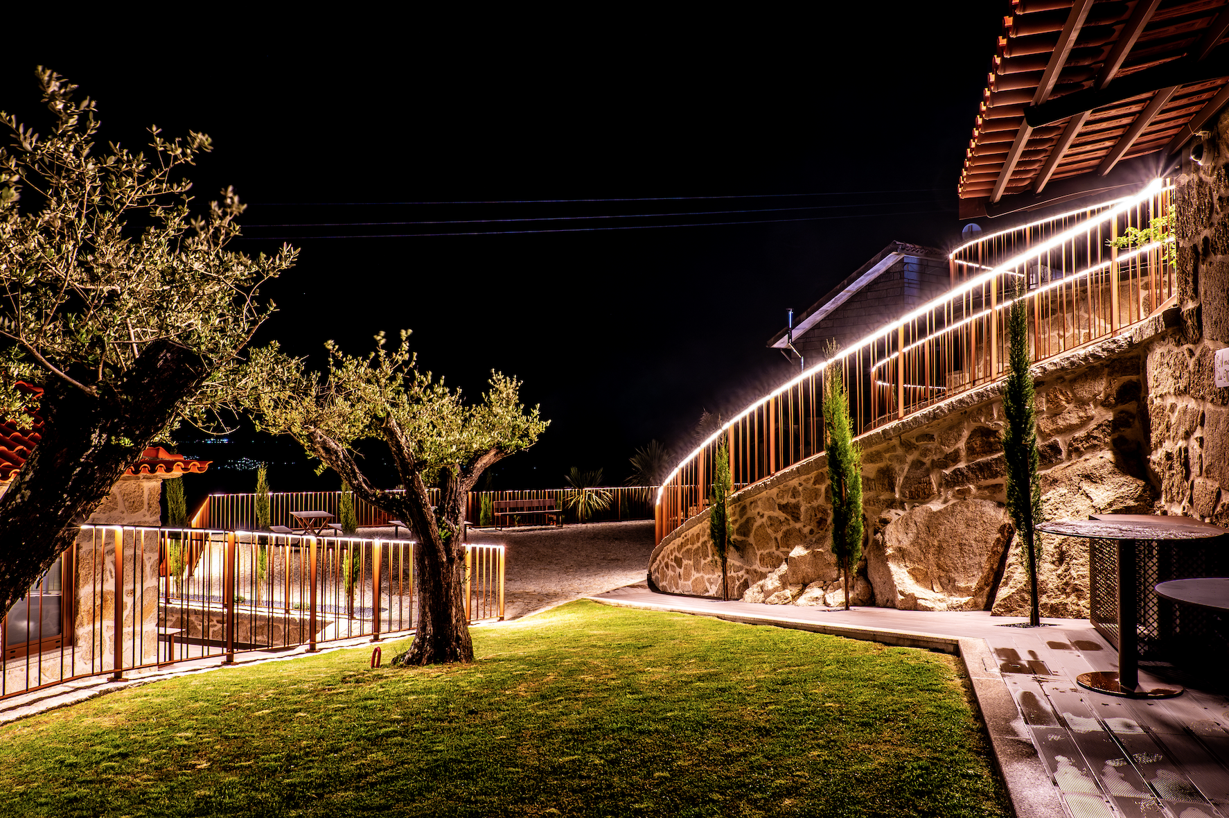 Cenário noturno com árvore, piso de grama, muro de pedra, piso de madeira, iluminação decorativa iluminando a cerca, árvores pequenas, bancos de madeira, e casas ao fundo.