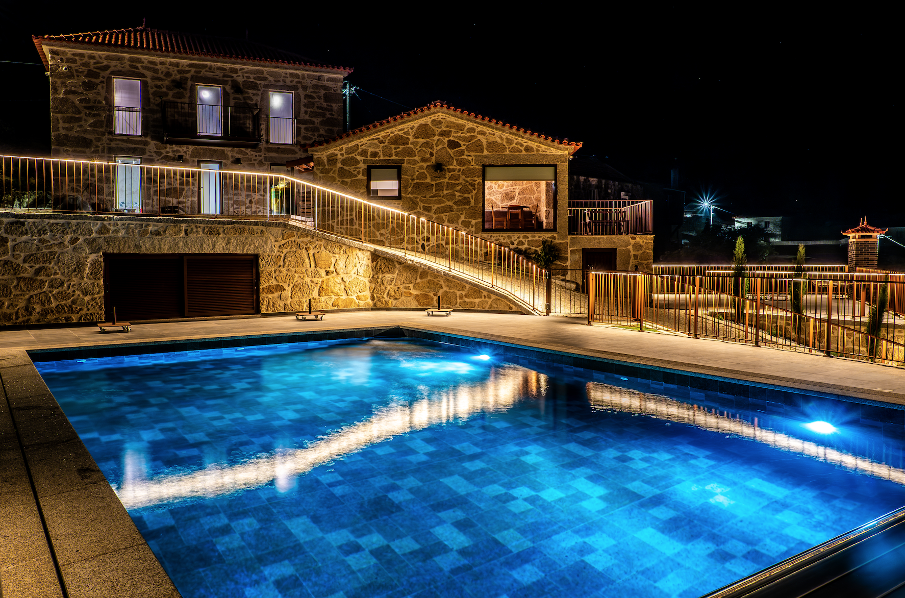 Casa de pedra iluminada à noite, com piscina de água azul na frente, cercada por guarda-corpos de ferro e iluminação decorativa