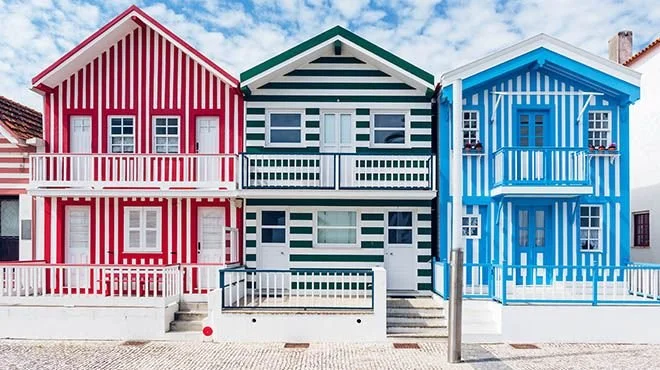 Três casas coloridas lado a lado: uma vermelha com listras brancas, uma verde com listras brancas e uma azul com listras brancas, todas com varandas frontais e cercas brancas.