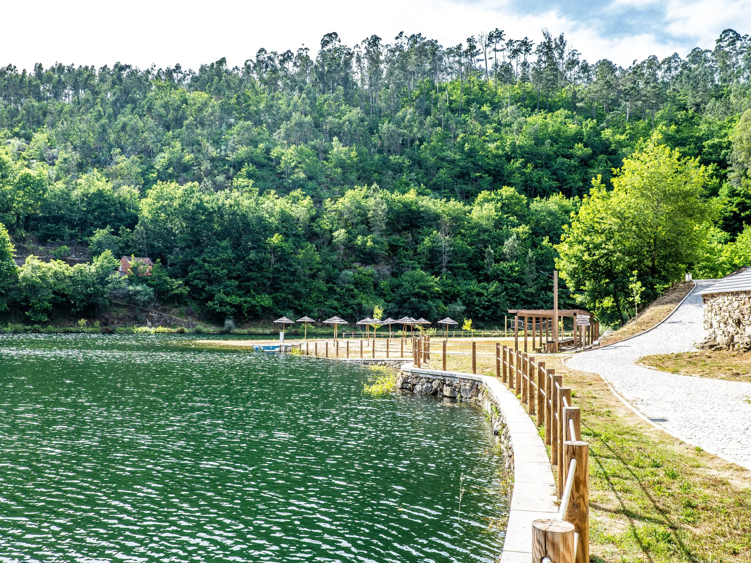 Cena de um rio sereno com água verde, ao lado de uma trilha de pedras com uma cerca de madeira, e uma floresta densa de árvores verdes ao fundo sob céu parcialmente nublado.