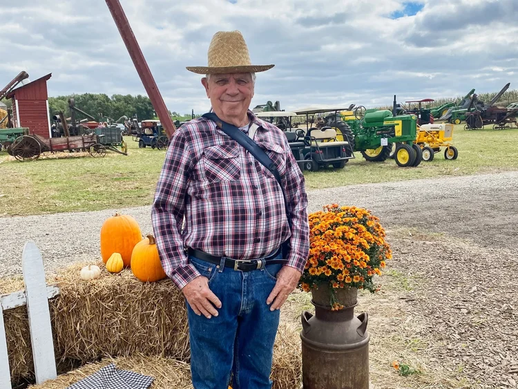 Galynn Ferris, founder and host of Tractorhead, in a straw hat and plaid shirt at an antique machinery show with restored tractors behind him
