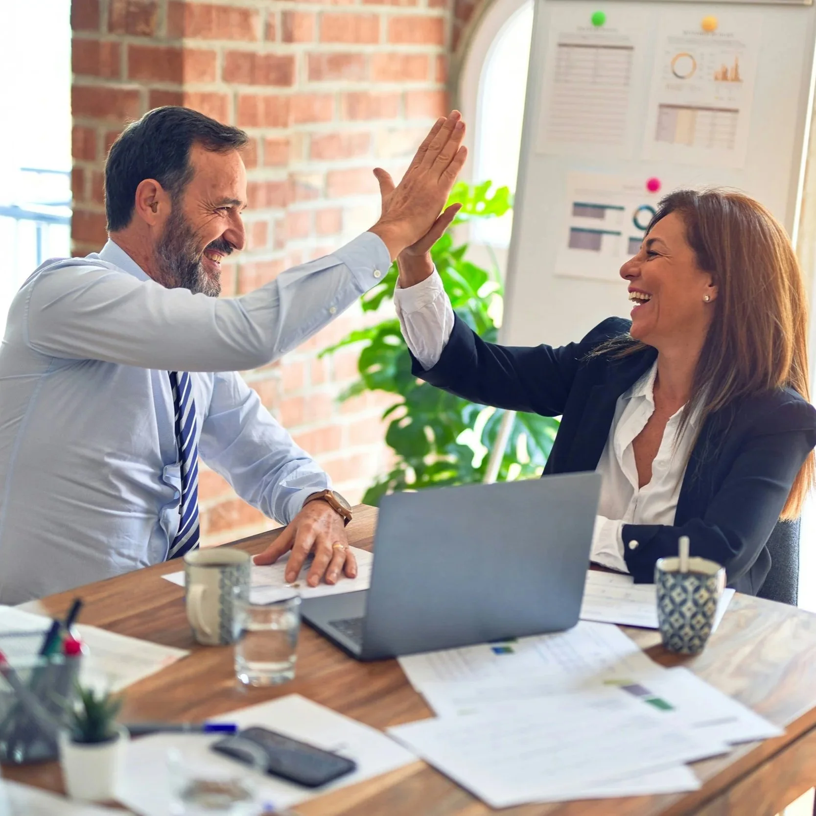 Two colleagues, a man and a woman, are giving each other a high five in a modern office. The man has a beard and is wearing a white shirt and a striped tie. The woman has shoulder-length brown hair and is dressed in a dark blazer over a white shirt. On the desk are a laptop, papers, pens, and mugs, with a brick wall and a window with blinds in the background.