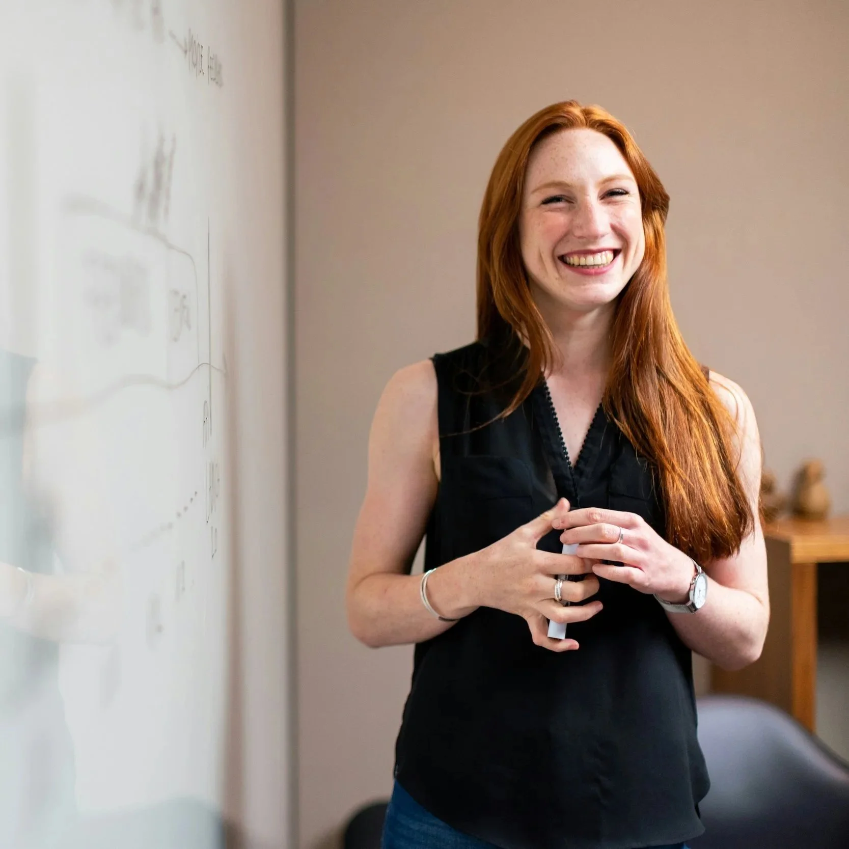 A woman with red hair and freckles wearing a sleeveless black top, holding a marker, smiling in front of a whiteboard.