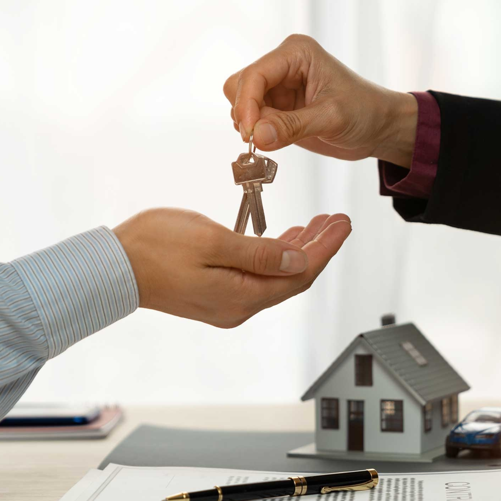Close-up of a hand holding a house key with a blank house-shaped keychain, with a desk, open documents, a pen, and small model houses in the background.