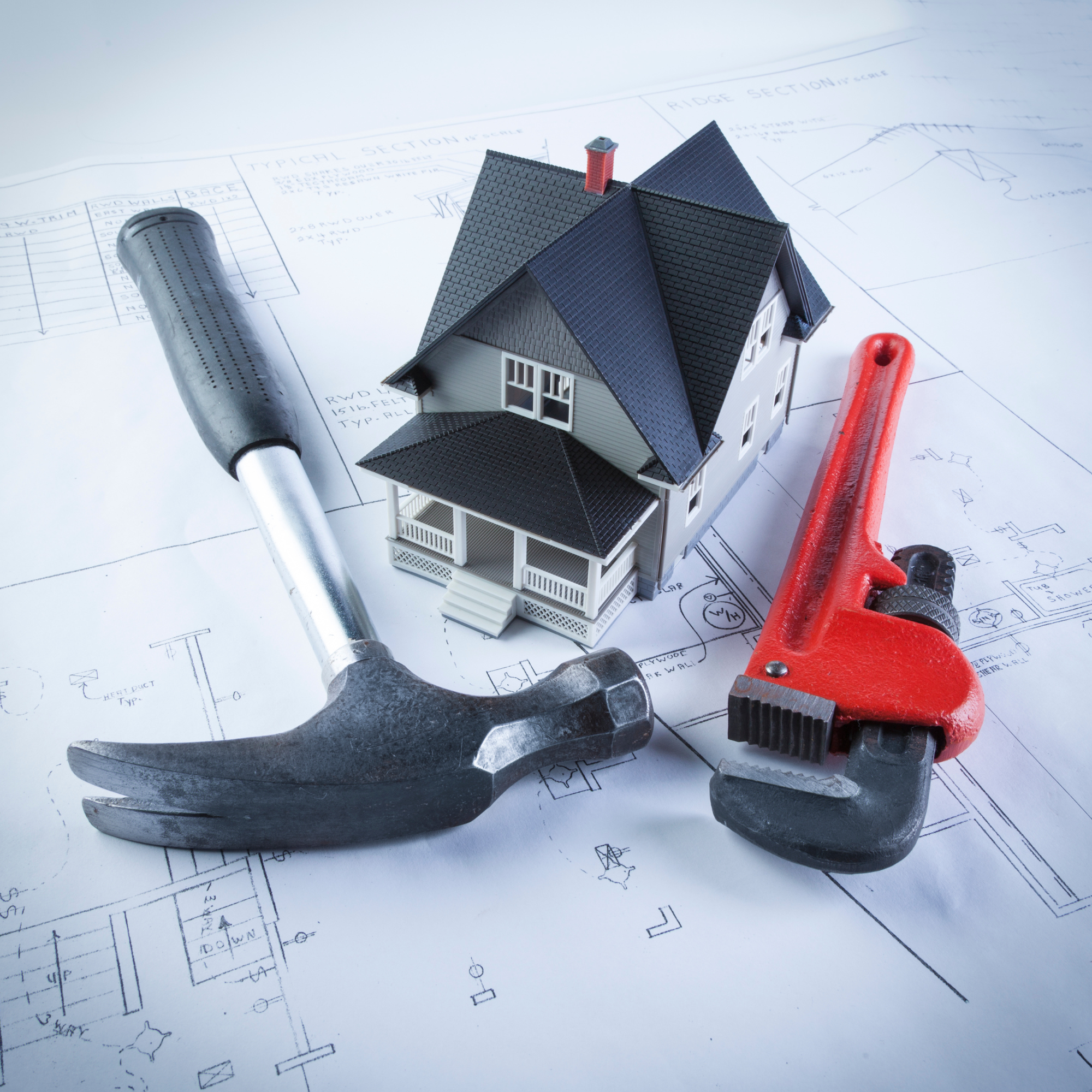 A model house on architectural blueprints with a black handle hammer, a silver pipe, and a red pipe wrench.