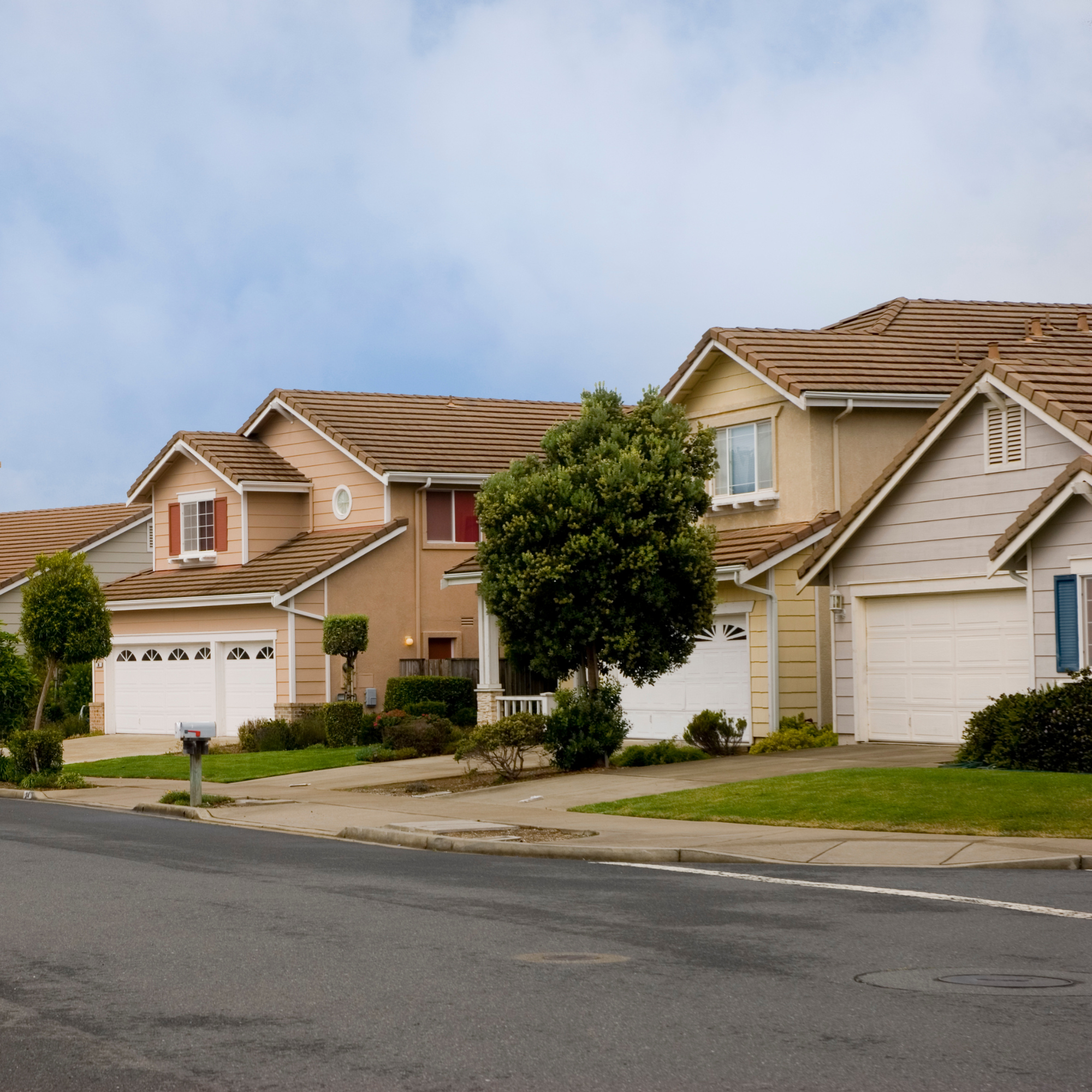 A suburban neighborhood with townhouses featuring beige and pastel colored exteriors, tiled roofs, and well-maintained yards with trees and shrubs. The street in the foreground is paved with a concrete sidewalk and a mailbox is visible. Overcast sky in the background.
