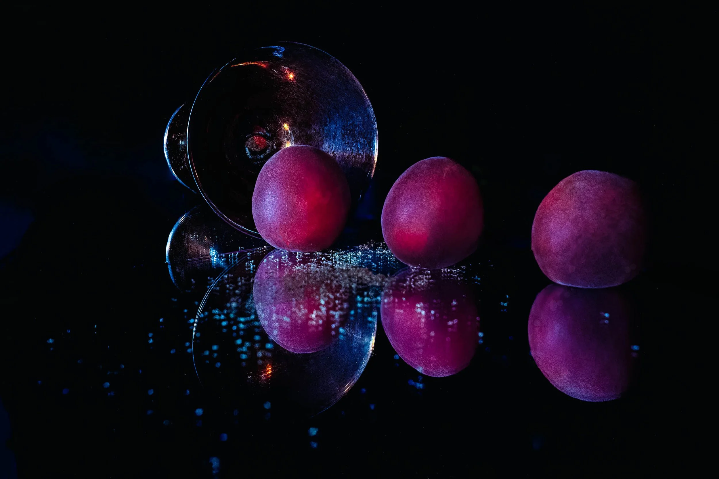 hree plums in a row beside a tipped wine glass, reflected on black glass in a dark studio still life