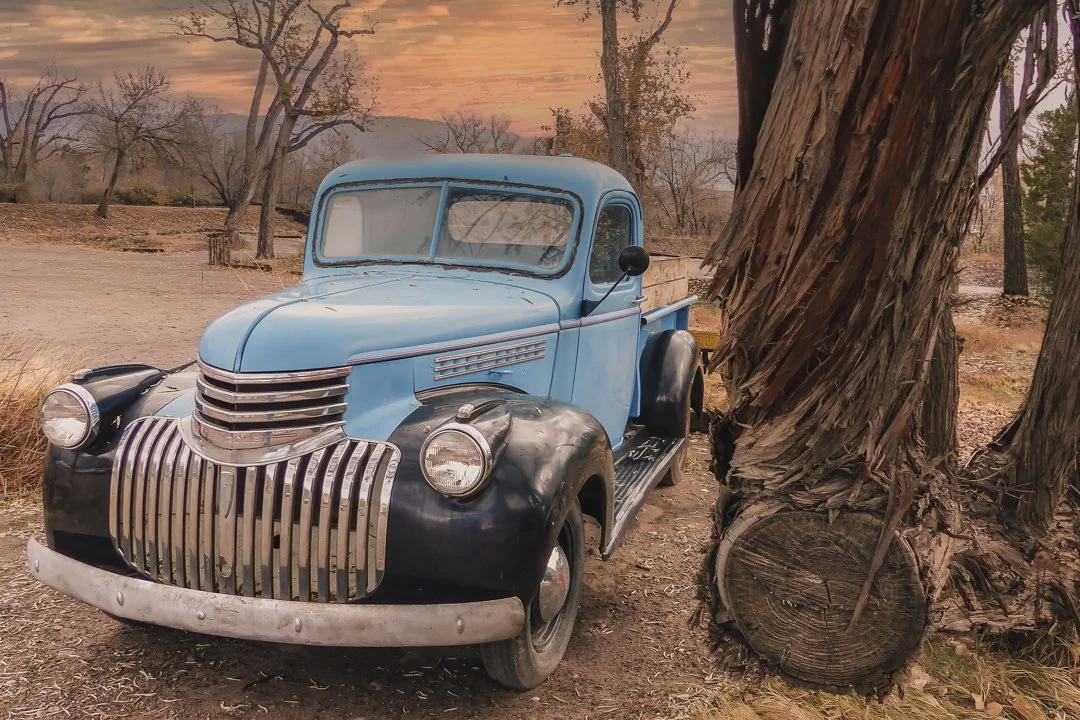 A faded vintage blue truck rests beneath towering cottonwood trees at dusk, weathered bark and soft evening light evoking quiet Southwest Americana