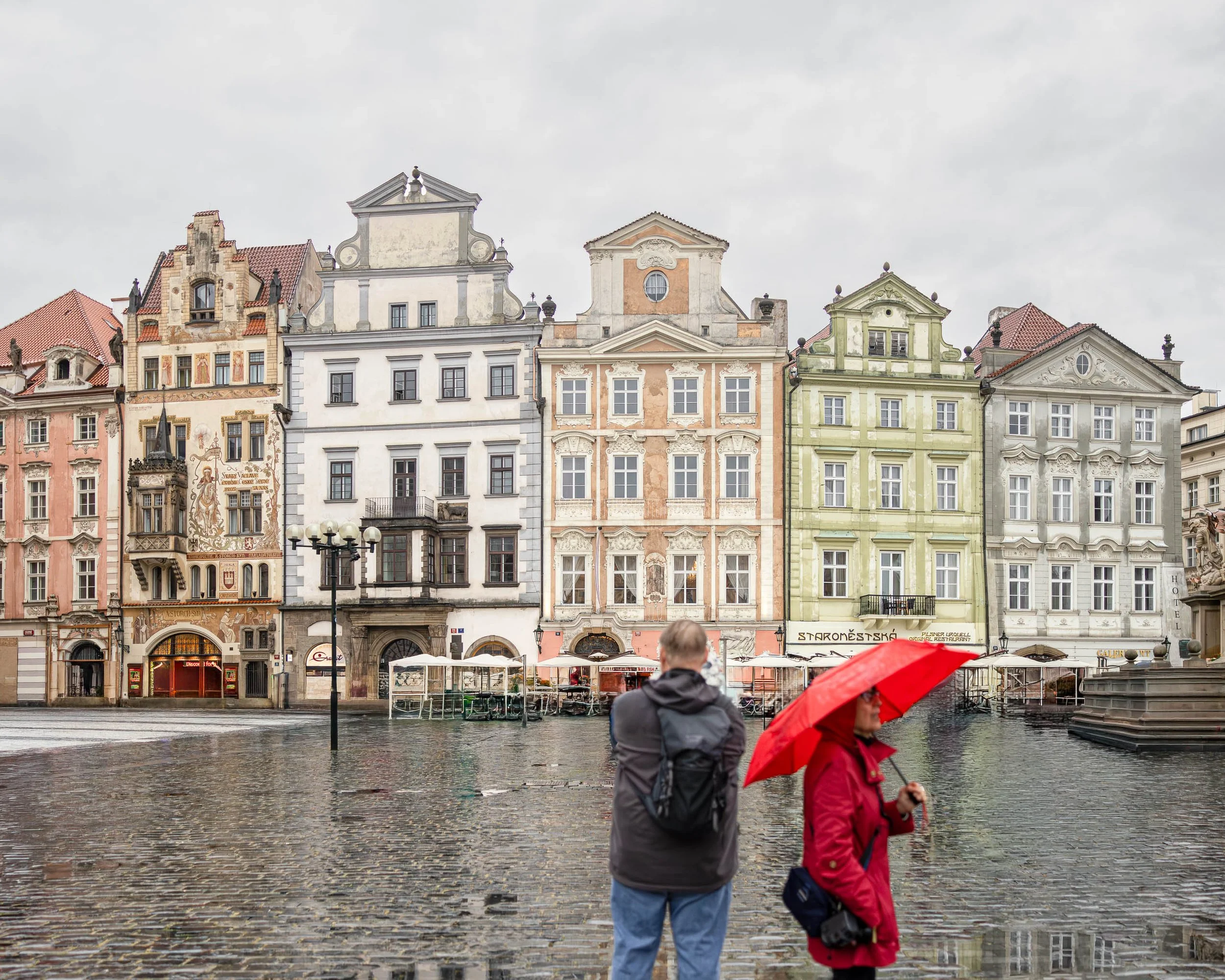 Two figures stand in Prague's Old Town Square during a rainstorm as historic pastel-colored buildings rise behind them and a red umbrella adds color to the scene.