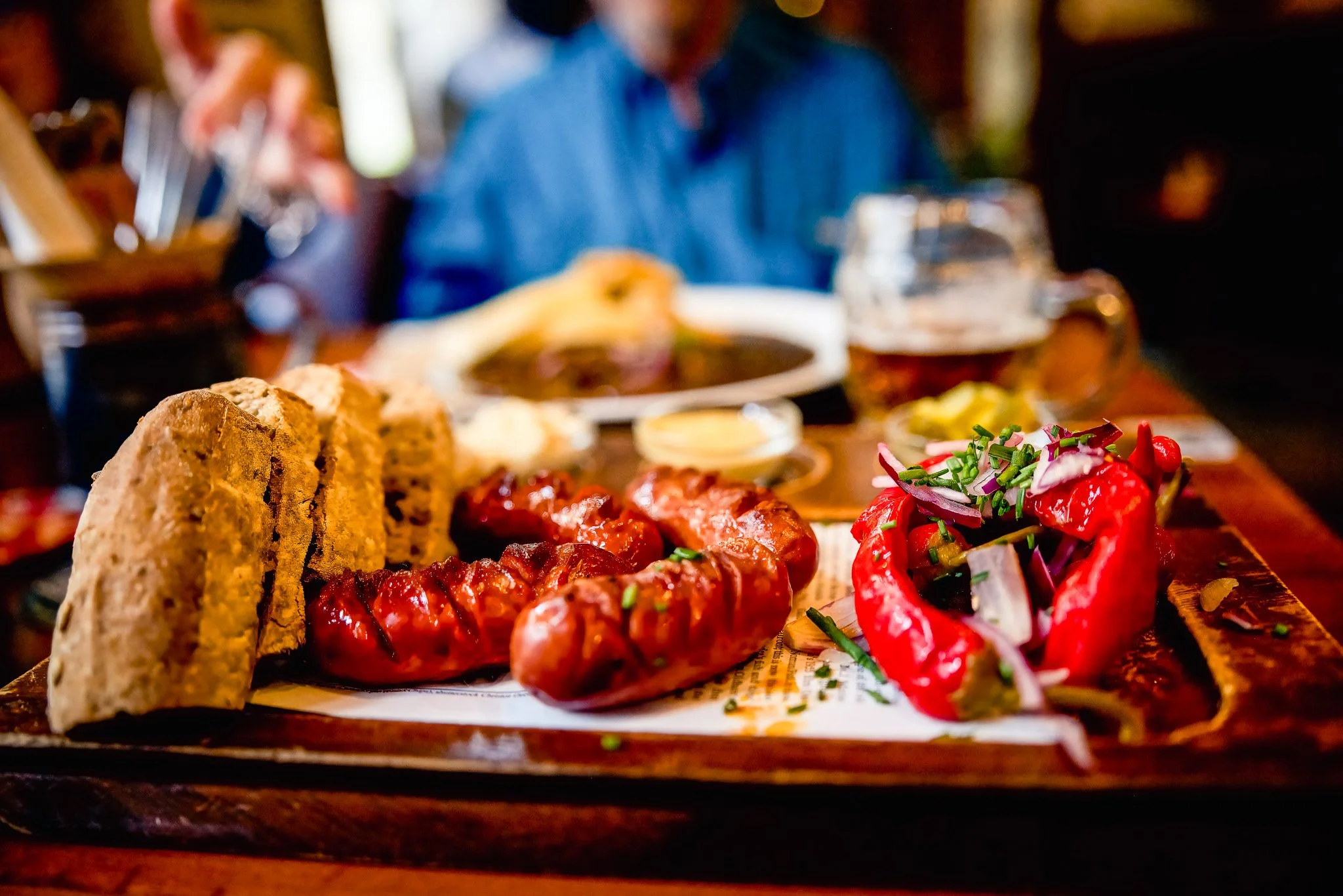 Traditional Prague lunch featuring grilled sausages, roasted peppers, rustic bread, and a softly blurred diner in the background.