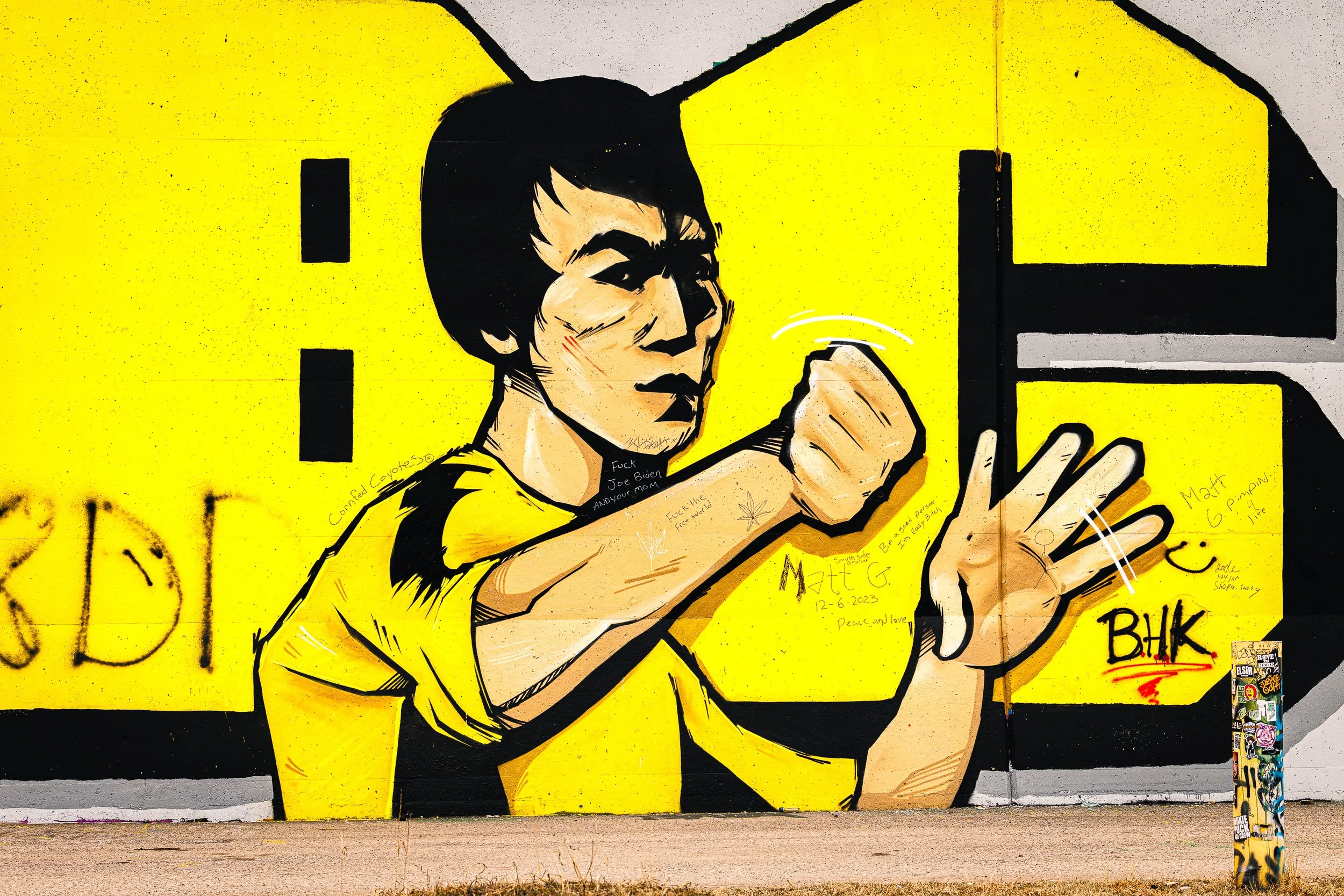 Street art mural of a woman with black hair in a yellow shirt, holding her fist up in a fighting pose, displayed on a yellow background with various tags and writings around.