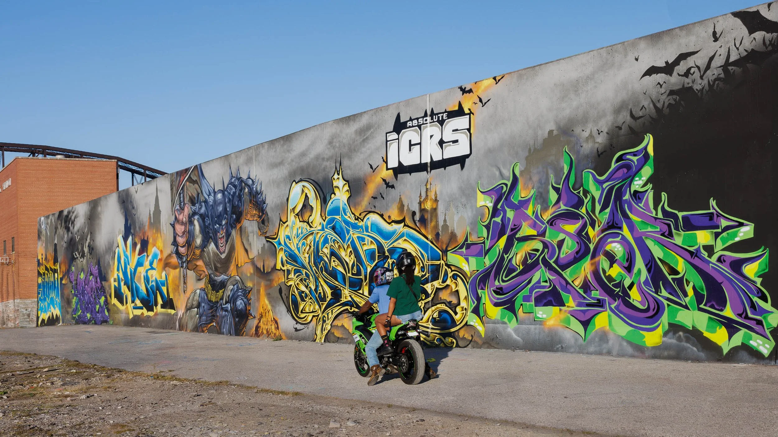 Two riders on a green motorcycle pass a long gray mural wall with gold, blue and violet-green lettering, St. Louis