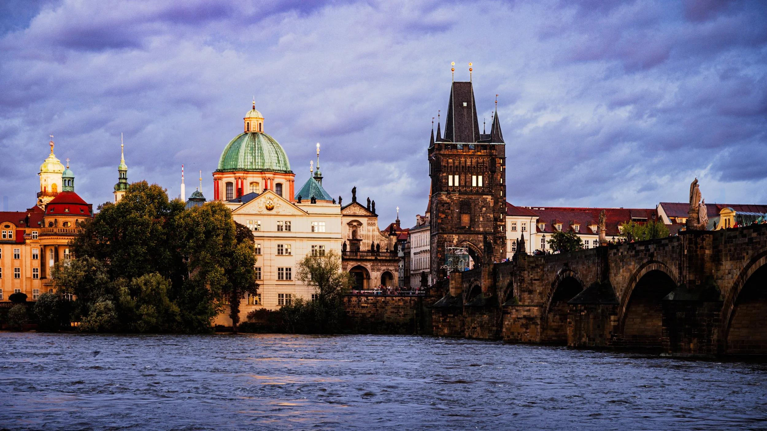 Storm clouds gather over Prague as warm evening light illuminates the Charles Bridge tower, historic domes, and buildings along the Vltava River.