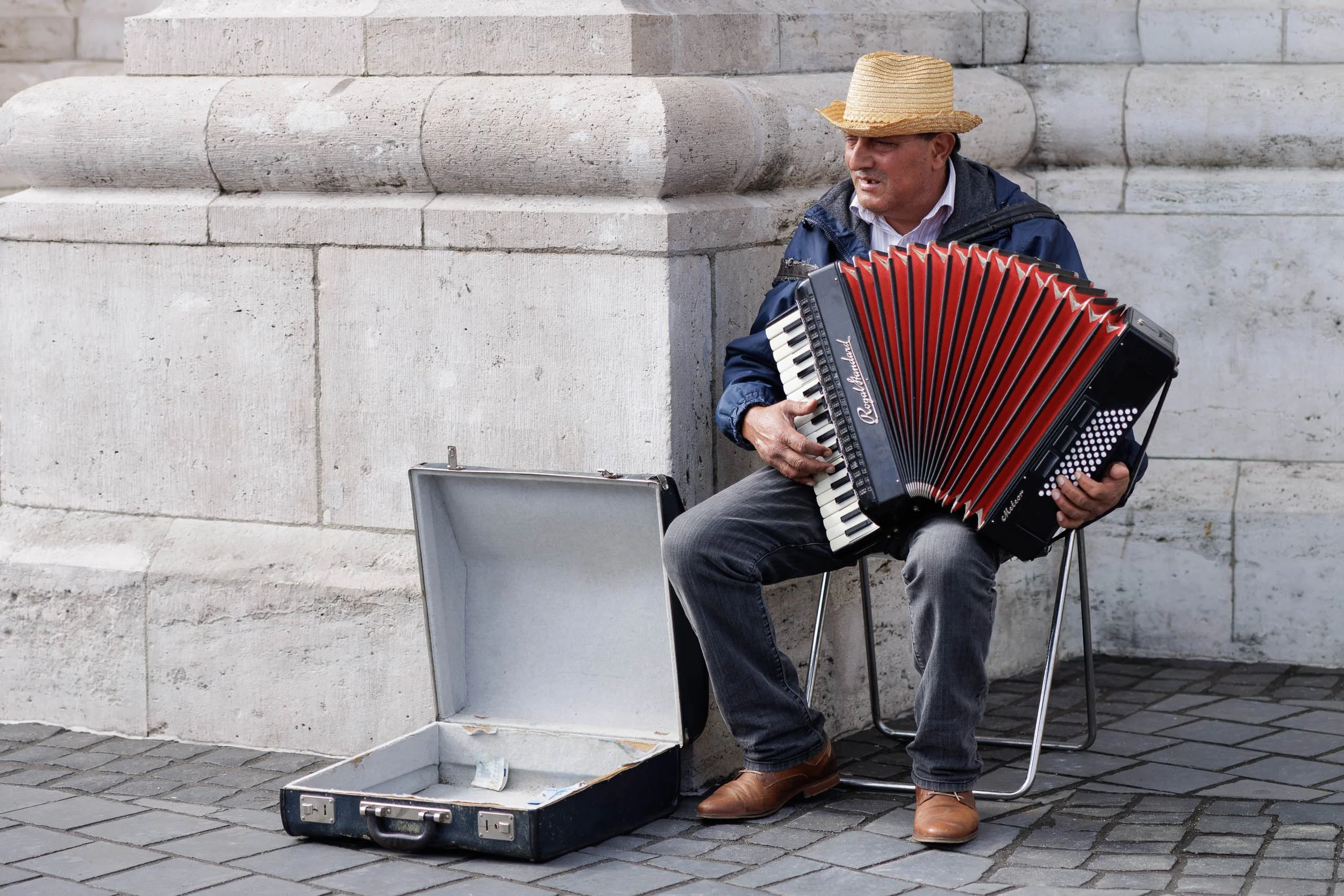 Street musician playing a red accordion beneath historic stone architecture in Budapest's Buda district, Hungary.