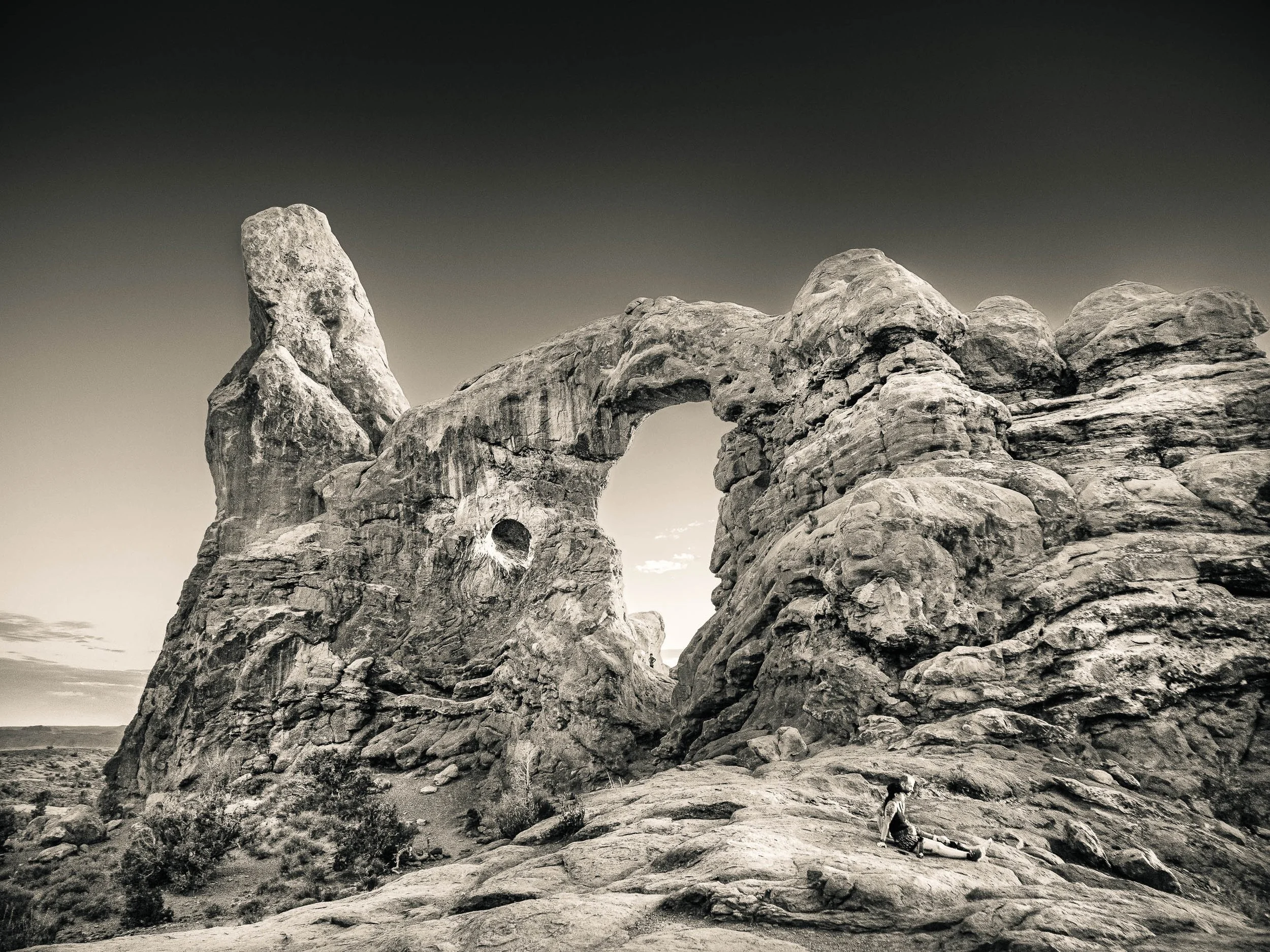A couple seated beneath a massive sandstone arch formation in Arches National Park, Utah, photographed in black and white.