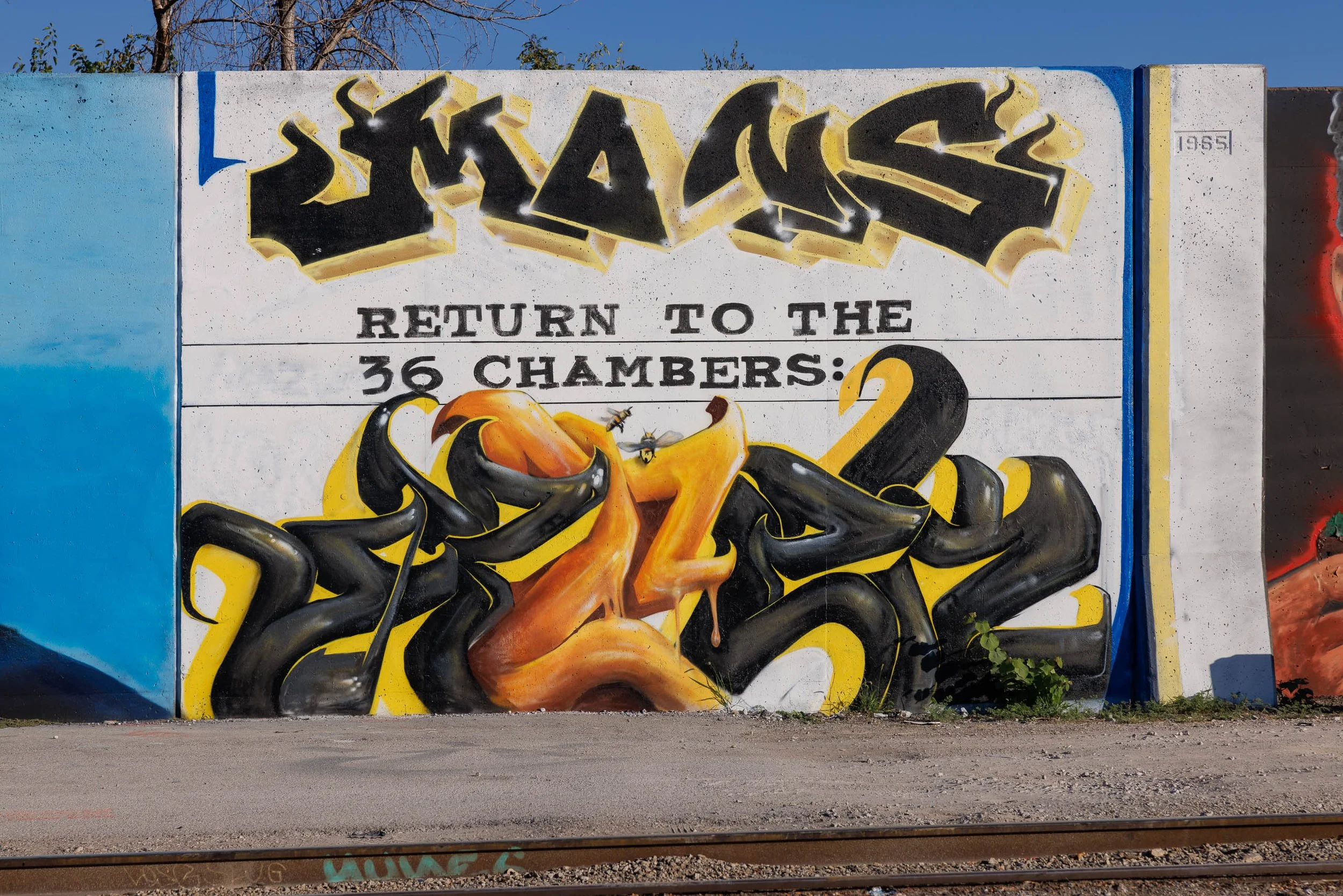 Black and honey-orange graffiti lettering on a white concrete wall in St. Louis with two bees hovering at the center beneath the words Return to the 36 Chambers