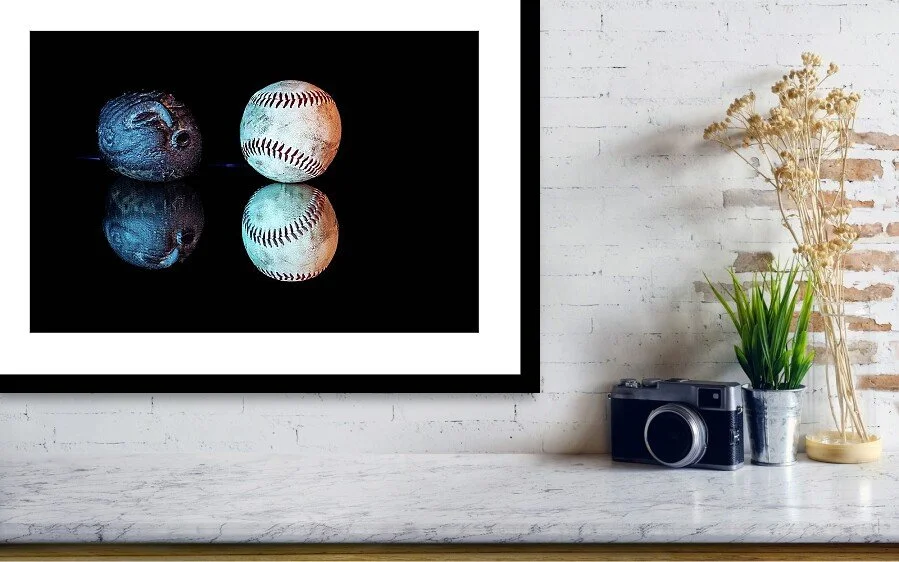 Color still-life photograph of a carved stone face appearing to blow toward a worn baseball, illuminated with blue and amber light and reflected against a black background.