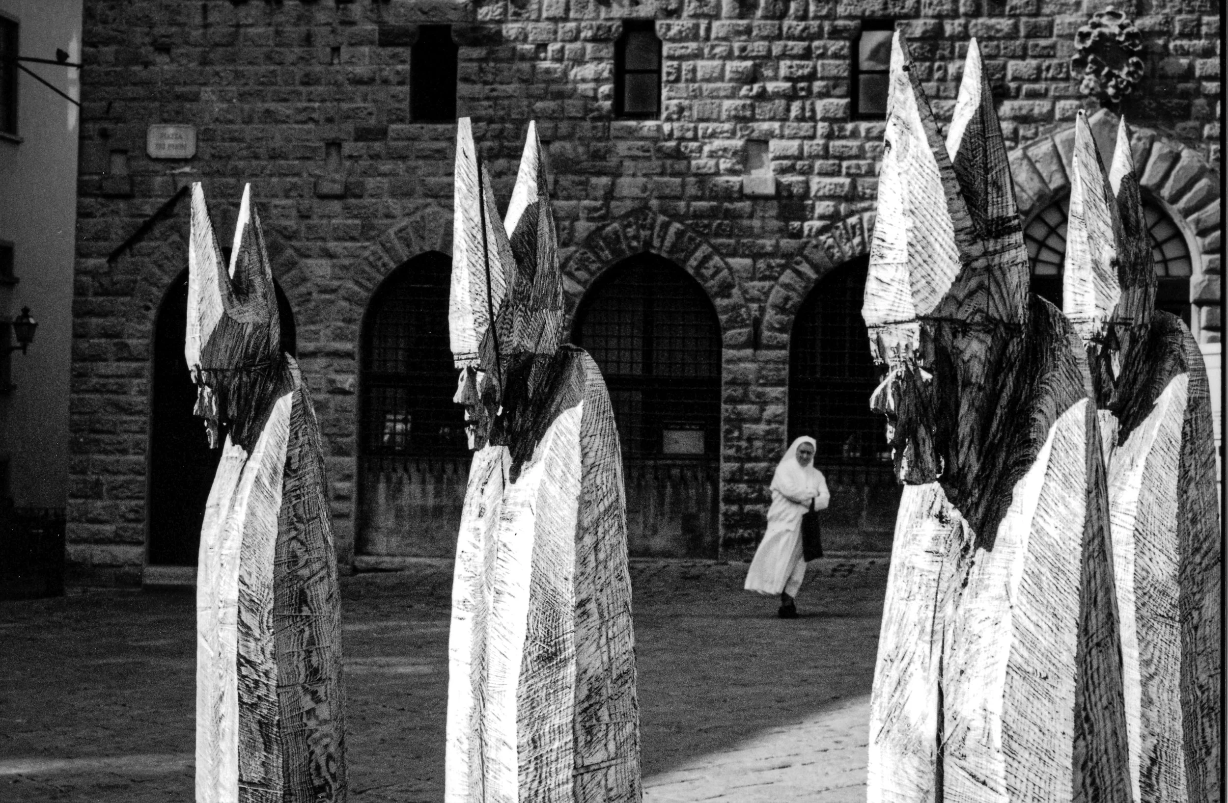 Black and white photo of a woman in a long dress walking among tall abstract wooden sculptures outside in front of a stone building with arched windows.