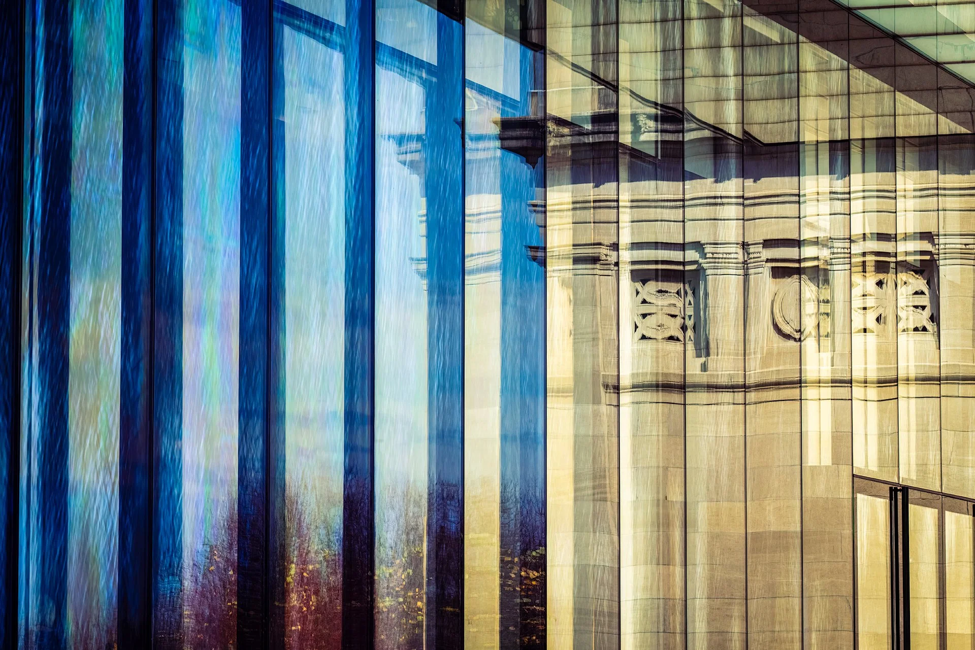 Reflected glass building with blue, beige, and green tinted windows, showing reflections of neighboring structures.