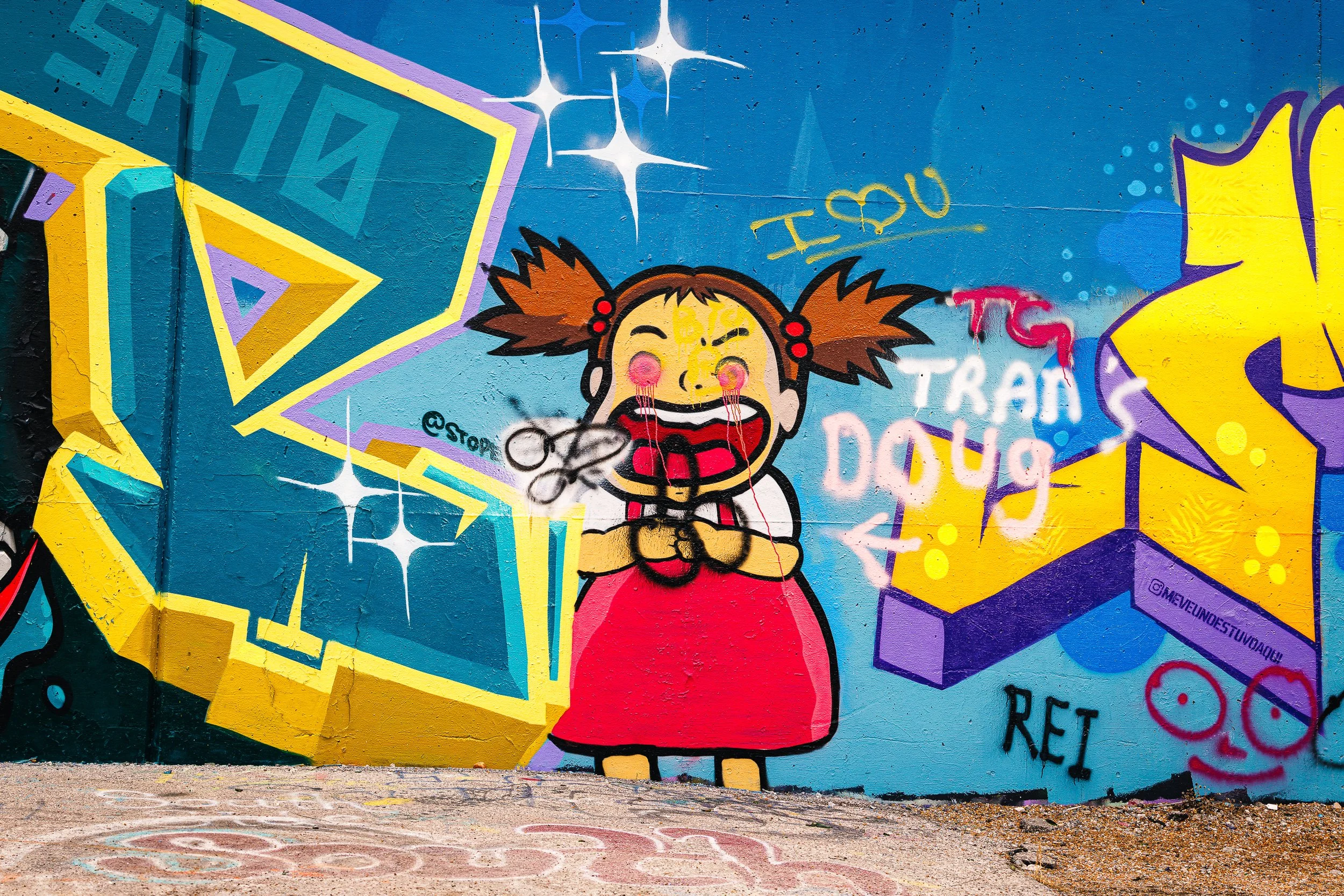 Colorful graffiti mural featuring a cartoon girl with pigtails, closed eyes, and blush on her cheeks, wearing a red and black striped shirt. The mural includes vibrant geometric shapes, stars, and text such as 'I ♥ U'.
