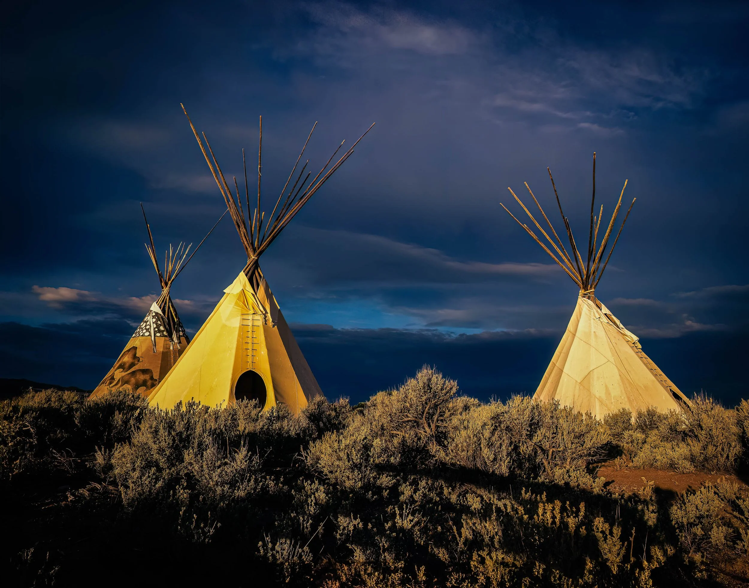 Three illuminated tipis beneath dramatic storm clouds in the American Southwest during a brief break in the weather.