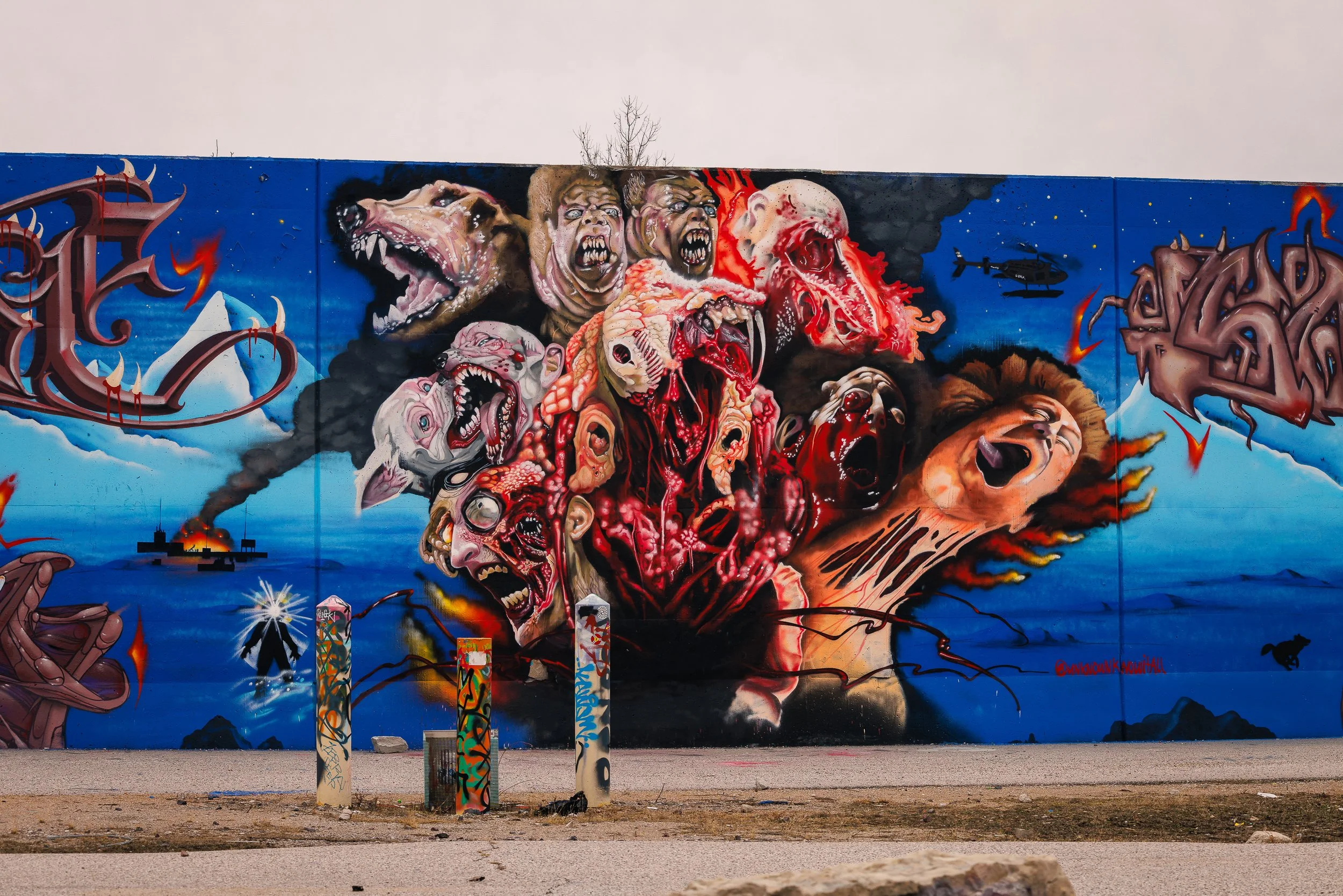 Colorful graffiti mural depicting a chaotic scene of various monstrous and zombie-like faces with open mouths, fangs, and distorted features, set against a blue background with mountains and clouds, and a helicopter silhouette.