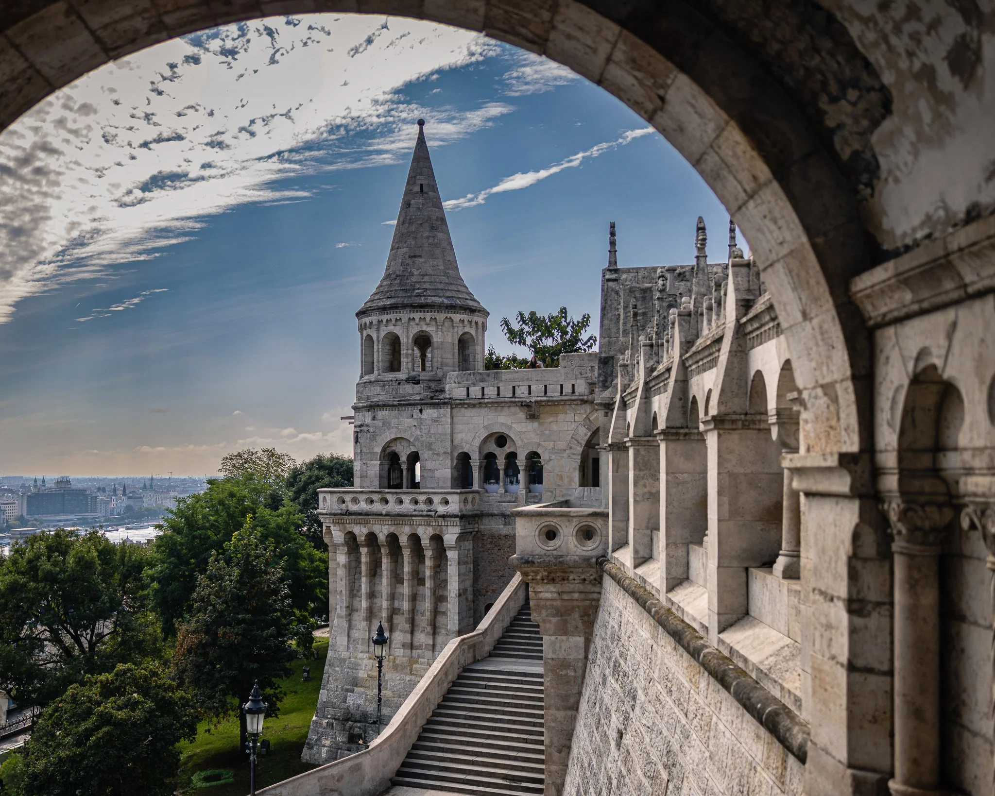 Stone archway framing Fisherman's Bastion tower and staircase in Budapest, Hungary beneath a dramatic blue sky.