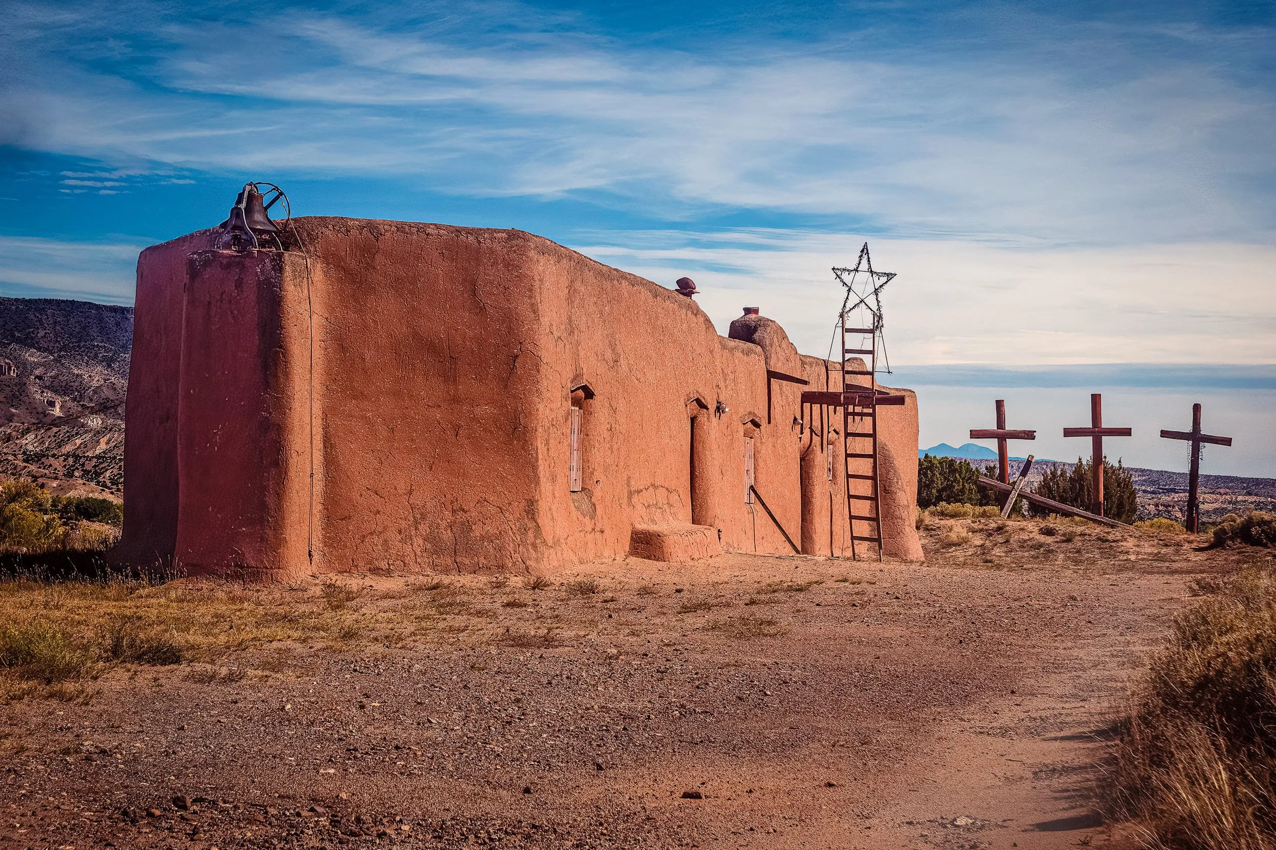 Historic adobe church near Abiquiú, New Mexico with bell, wrought-iron star, and three wooden crosses in the distance — fine art photography by Robert Niemeier