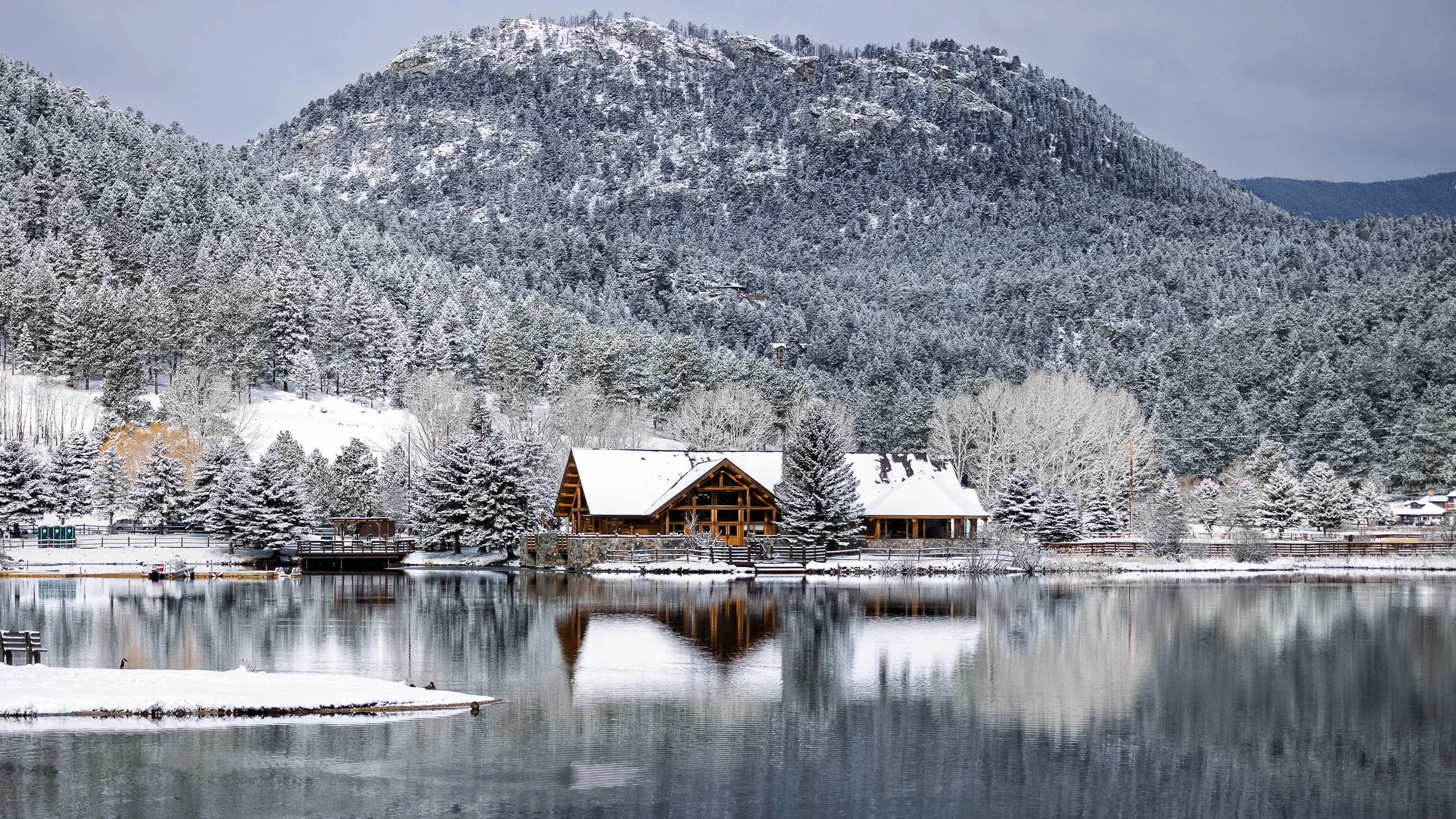 Fine art landscape photograph titled Where Winter Waited, featuring a snow-covered cabin and pine trees perfectly reflected in a still mountain lake with an alpine peak in the background.