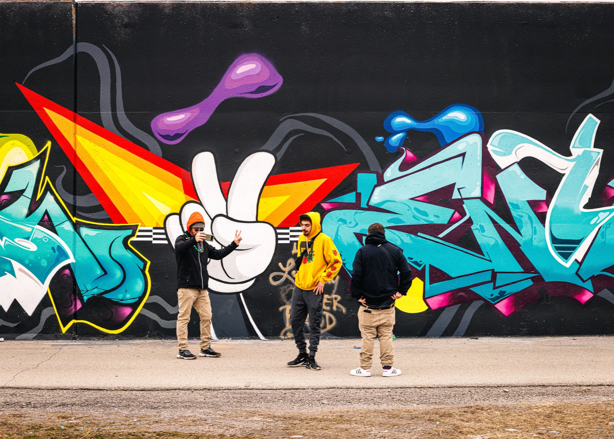 People standing in front of a vibrant graffiti mural on a black wall, with two individuals posing and another person holding a phone. The graffiti features colorful abstract designs and a large pair of cartoonish eyes.