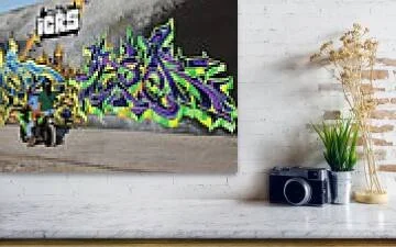 Two riders on a green motorcycle pass a long gray mural wall with gold, blue and violet-green lettering, St. Louis shown partially on mantle