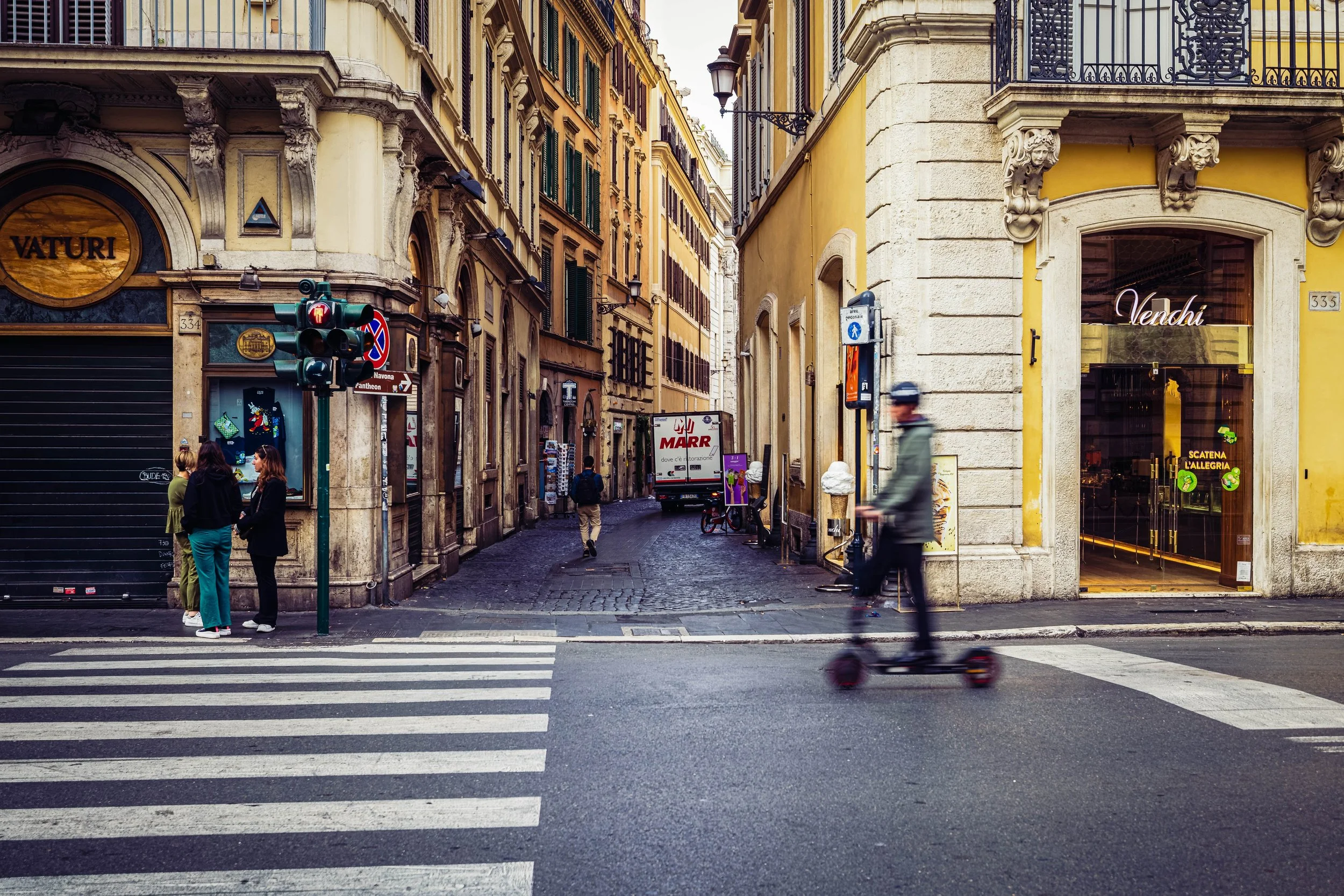 Candid street scene in Rome showing a blurred scooter rider, pedestrians, and warm ochre building facades near a busy intersection.