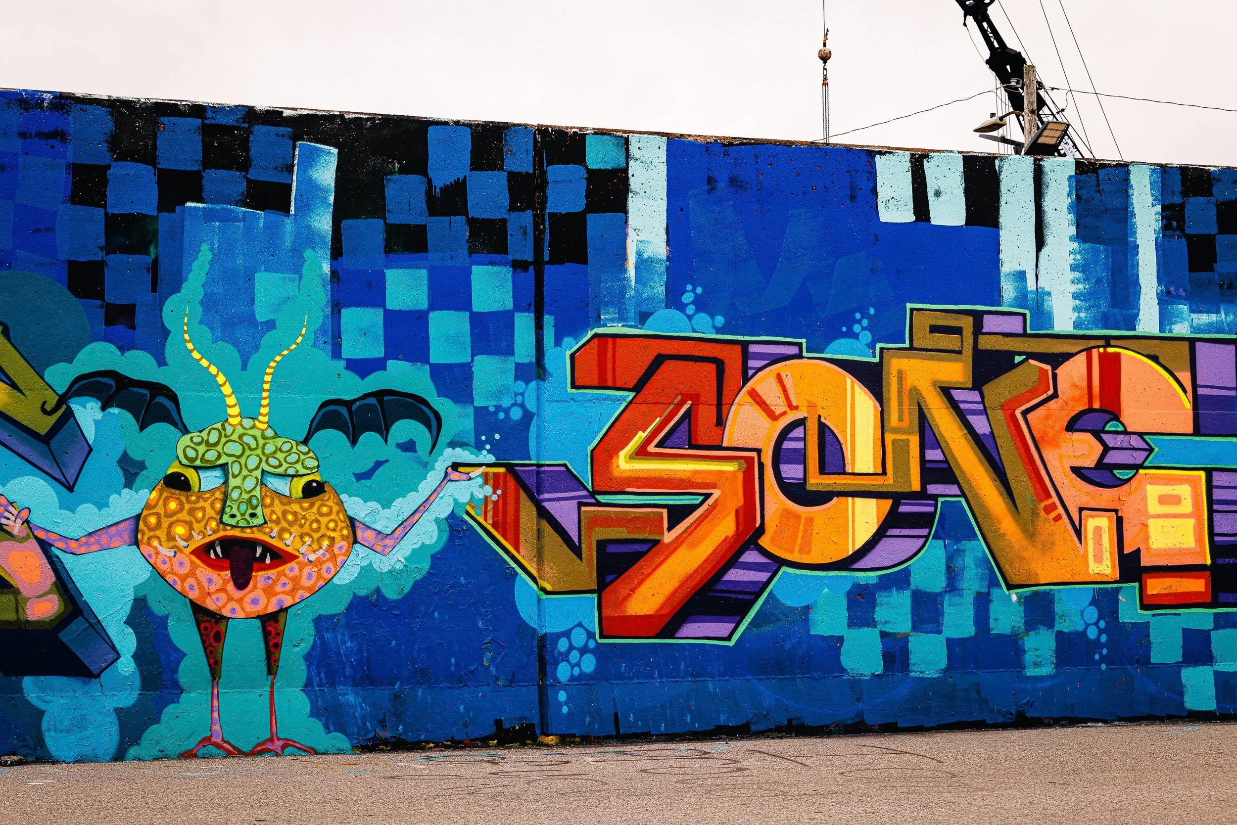 Colorful graffiti mural featuring a cartoonish creature with antennas and a human face with an open mouth, along with abstract geometric shapes and checkered patterns.