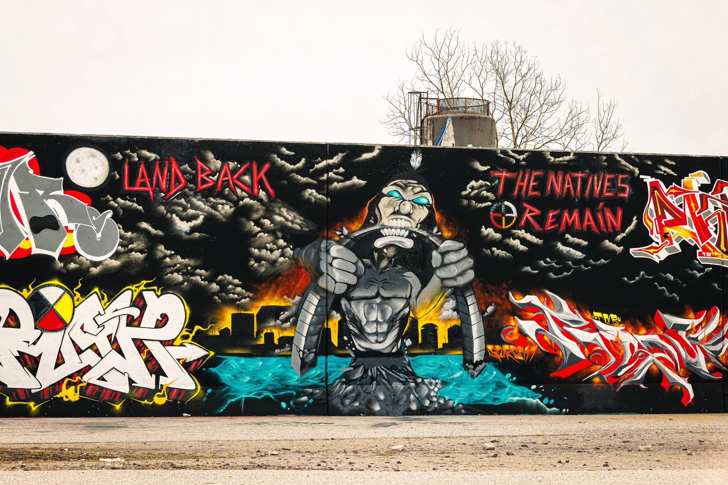 Colorful graffiti mural featuring a muscular figure with glowing blue eyes, surrounded by dark clouds, a city skyline, and bold text that says 'Land Back' and 'The Natives Remain.'